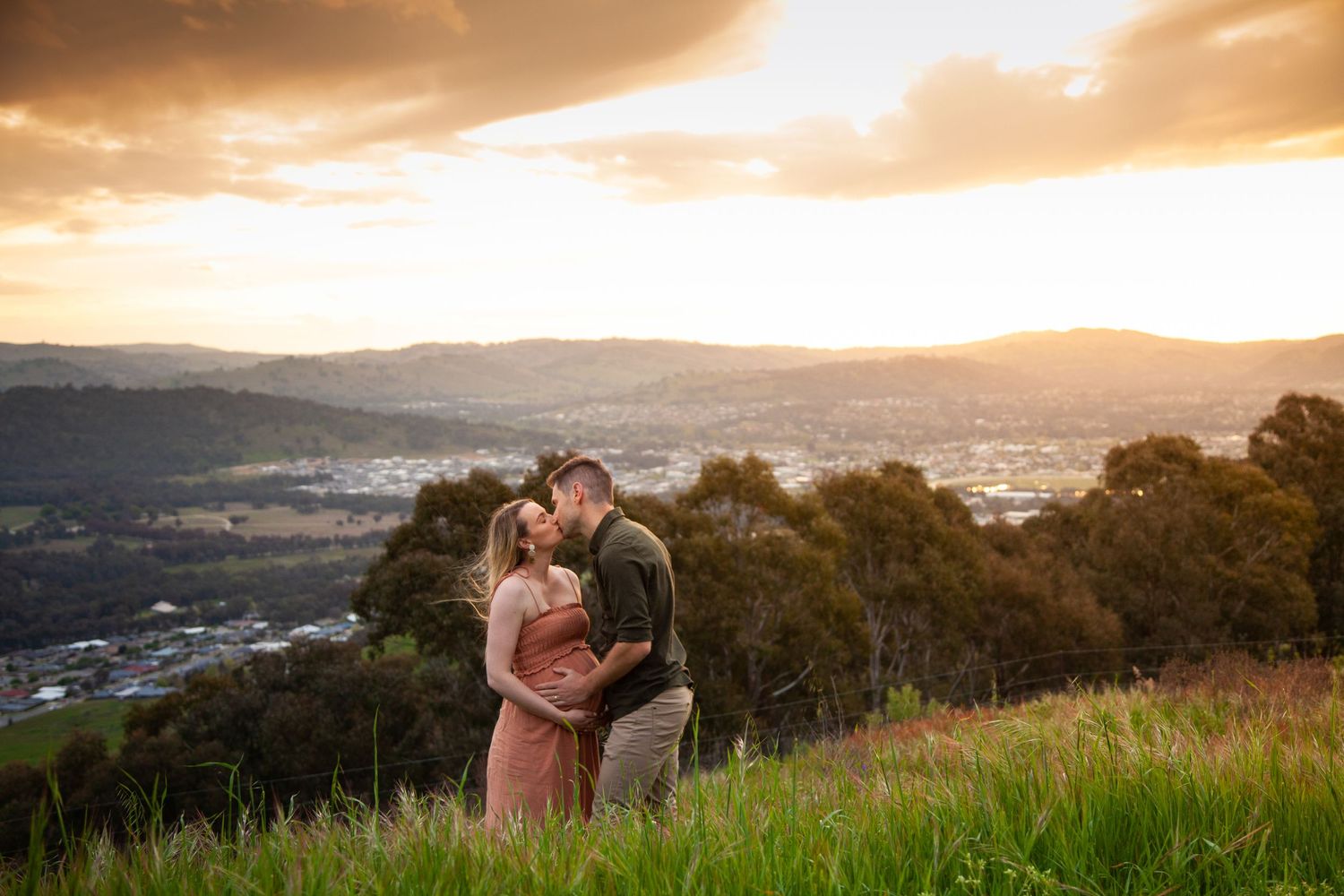 Romantic sunset maternity photo of a couple embracing on a hilltop overlooking a valley.
