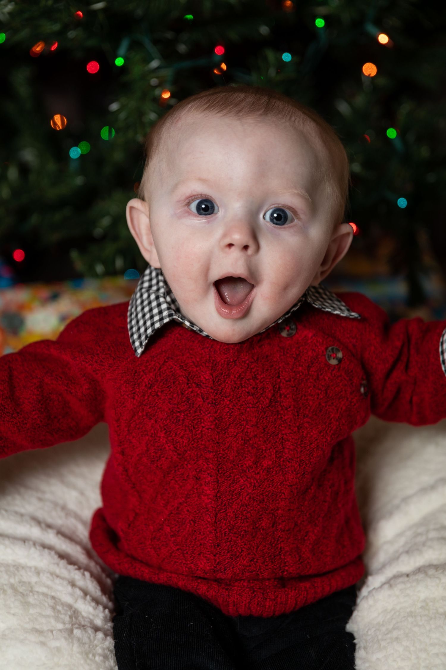 Baby in red sweater with checkered collar against Christmas lights background.