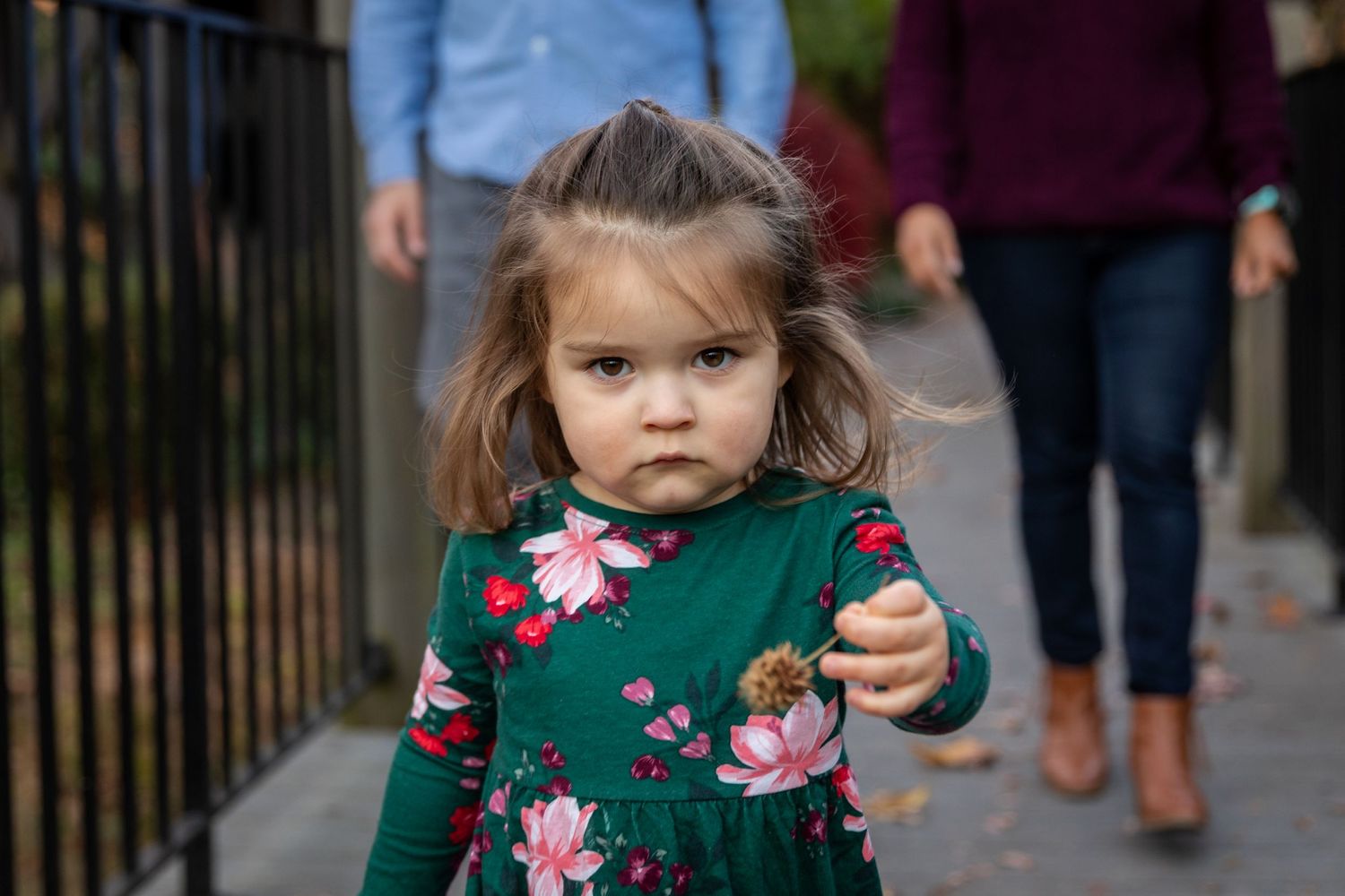 Young child wearing floral dress and boots walking along city sidewalk during fall.