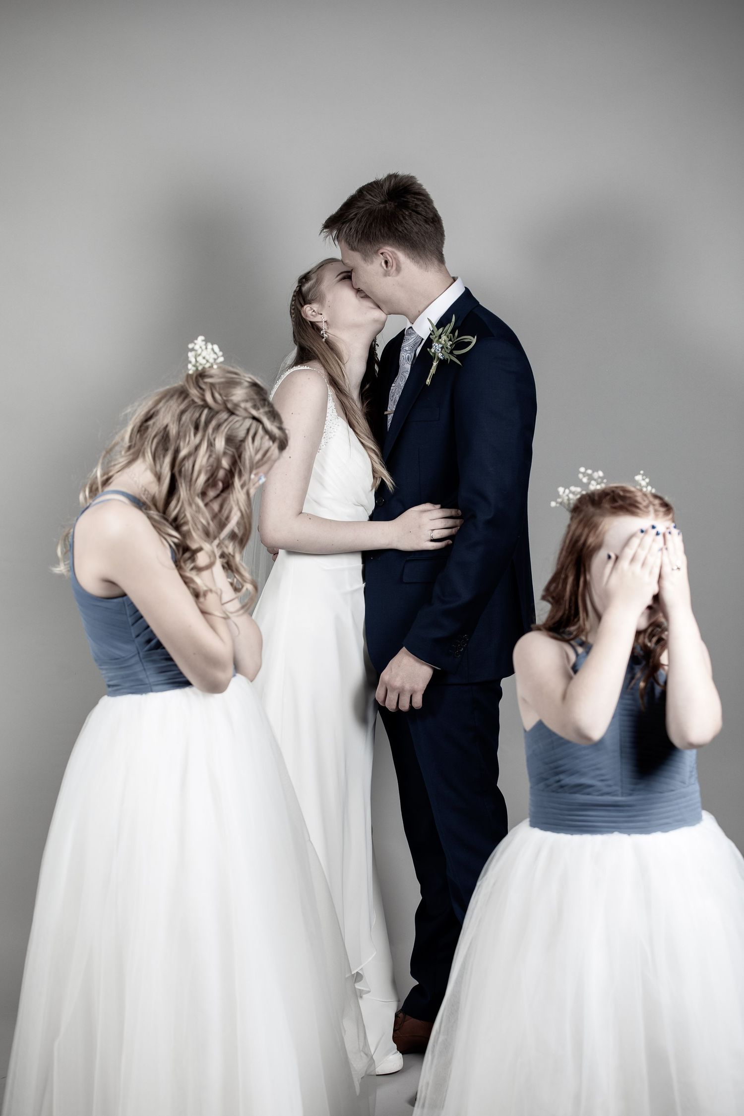 Wedding couple sharing a kiss while two flower girls cover their eyes in a playful studio portrait.