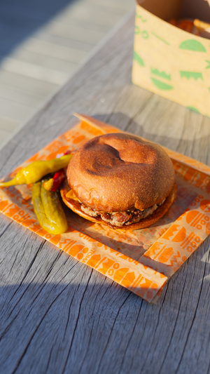 A close-up of a burger with pickles on orange patterned paper in natural sunlight.