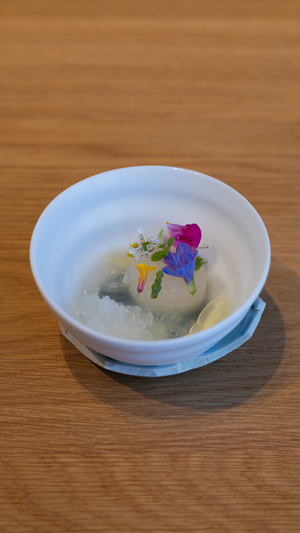 A white ceramic bowl containing a clear broth garnished with colorful edible flowers.