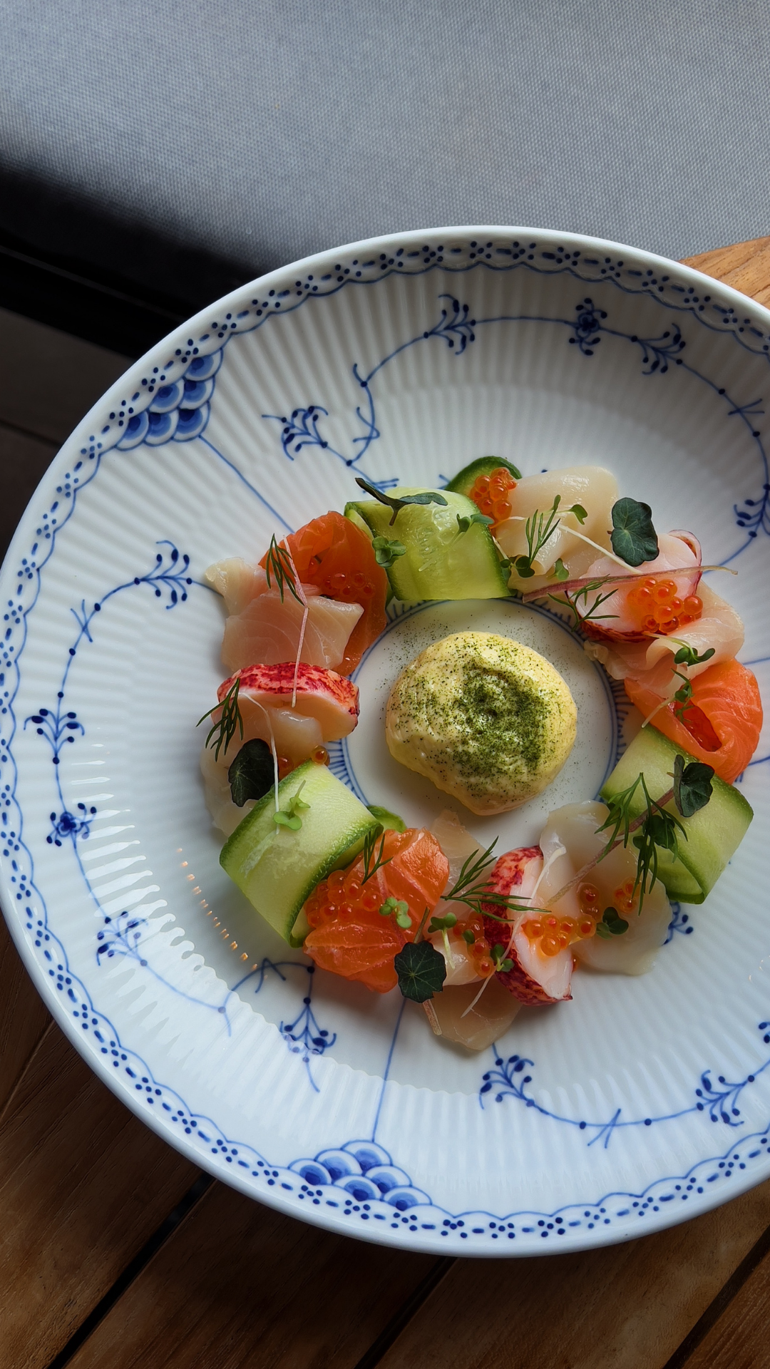 Elegant plating of cured fish and vegetables arranged in a circle on a blue and white porcelain plate with herb garnish.