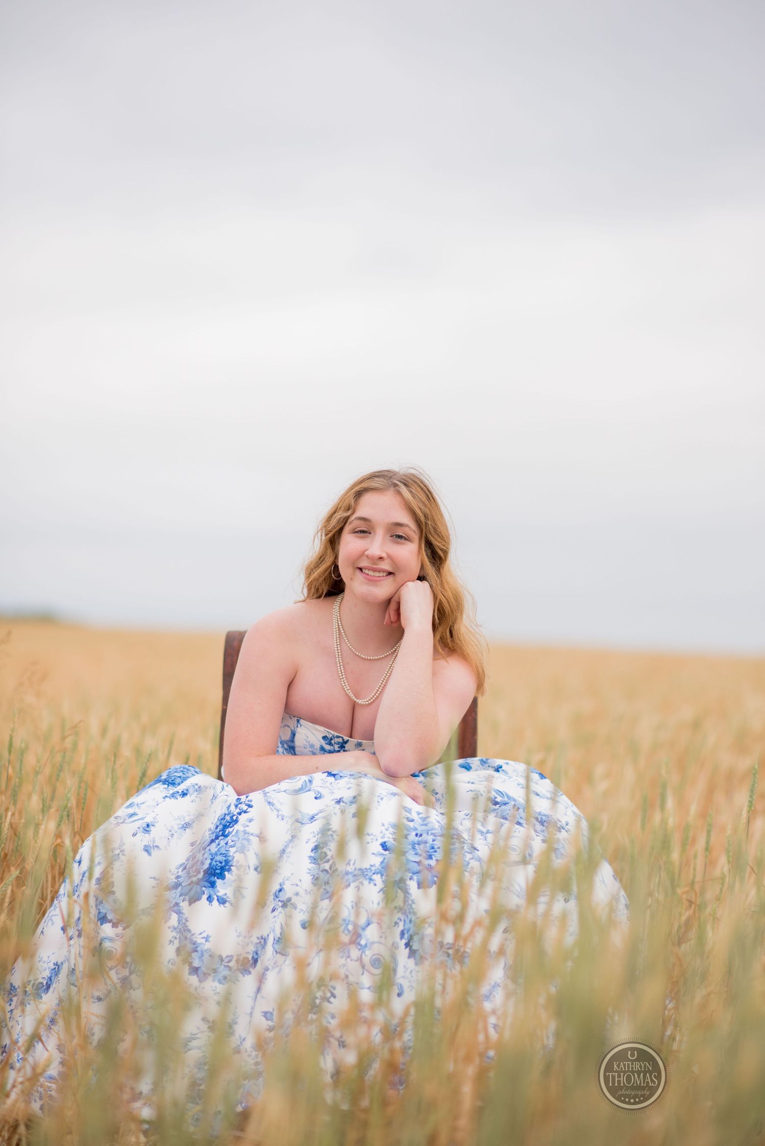 Portrait of a woman in a blue floral gown sitting in a brown chair surrounded by golden wheat.
