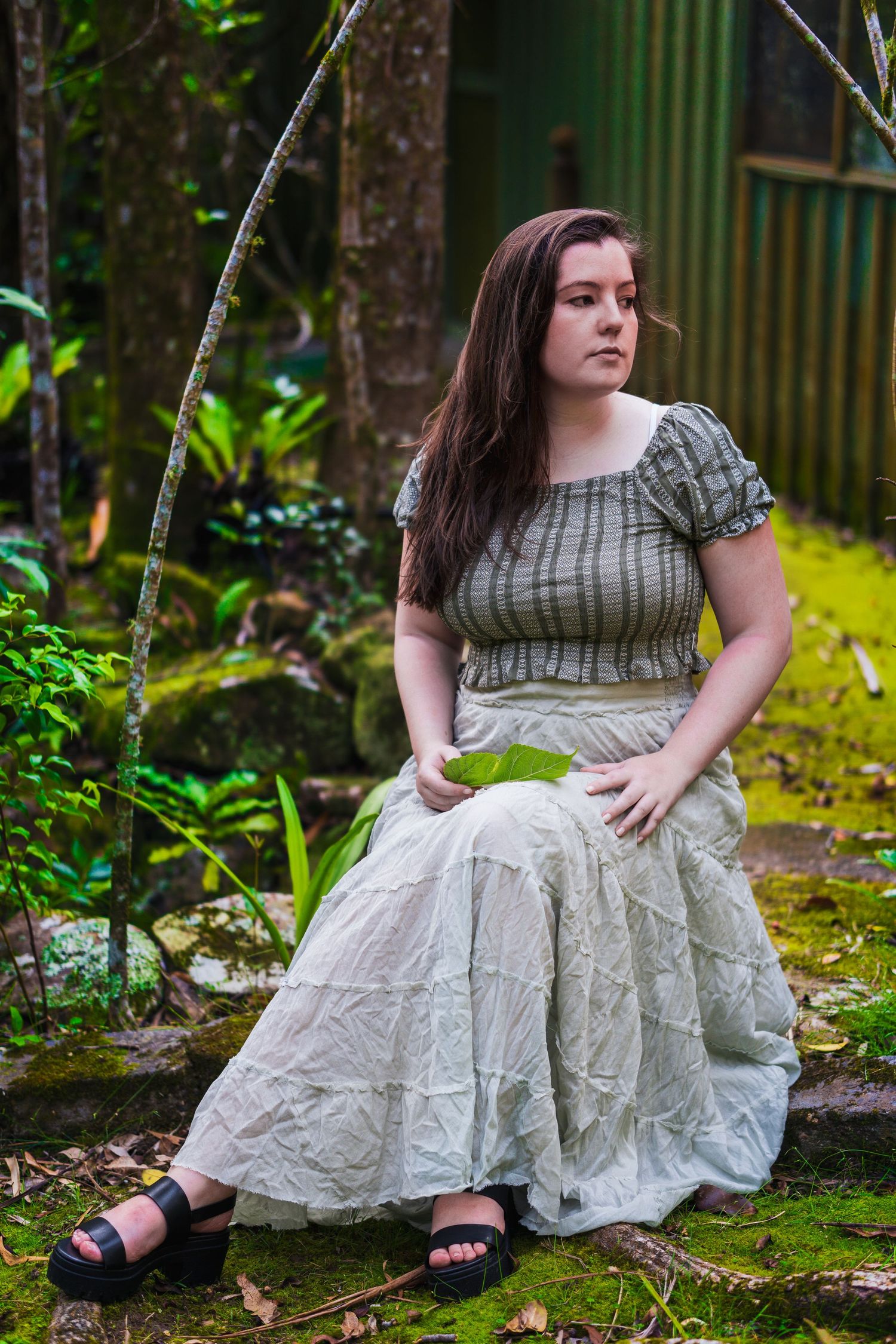 Person in vintage-style dress sits thoughtfully in a lush garden setting with moss and bamboo.