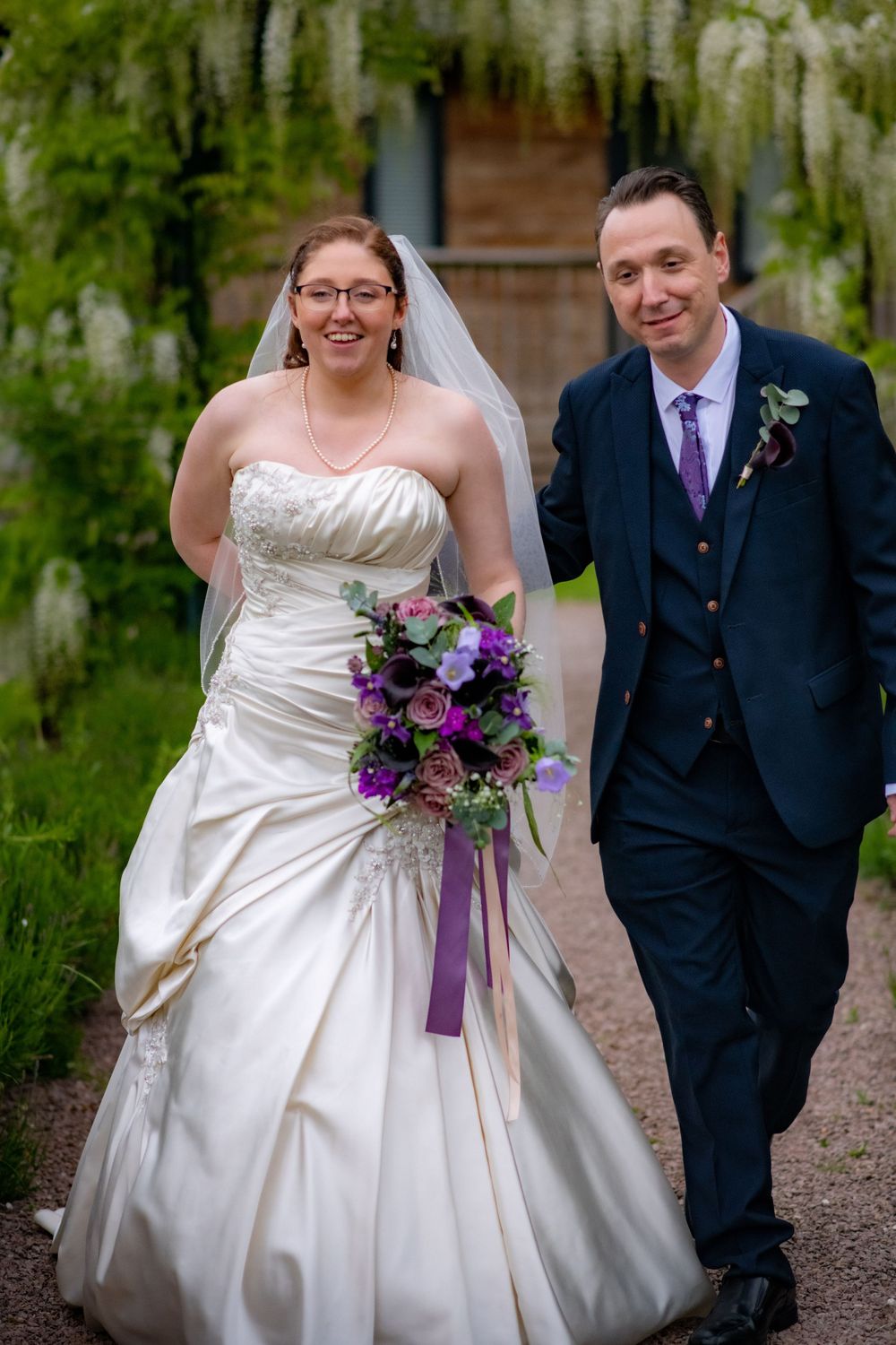 Newlyweds walking together with purple bouquet under wisteria covered walkway.