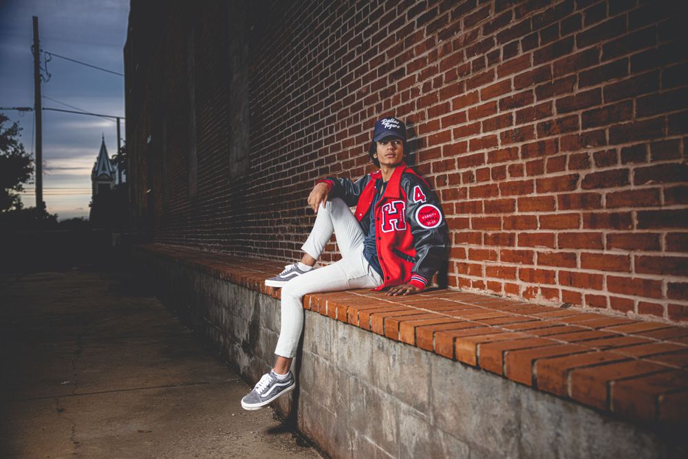 Baseball athlete in white pants and red jacket sits on brick ledge at dusk.