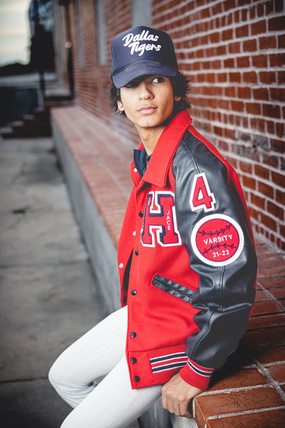 Youth in varsity jacket and baseball cap poses against brick wall wearing Dallas Tigers gear.