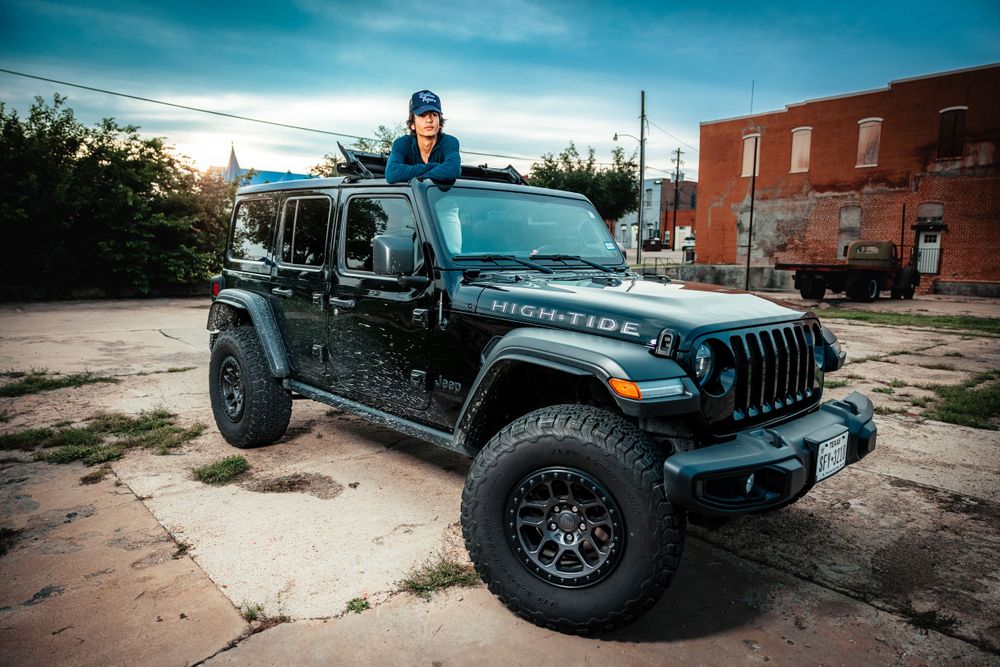 Person stands on top of black Jeep Wrangler with custom wheels parked in urban setting.