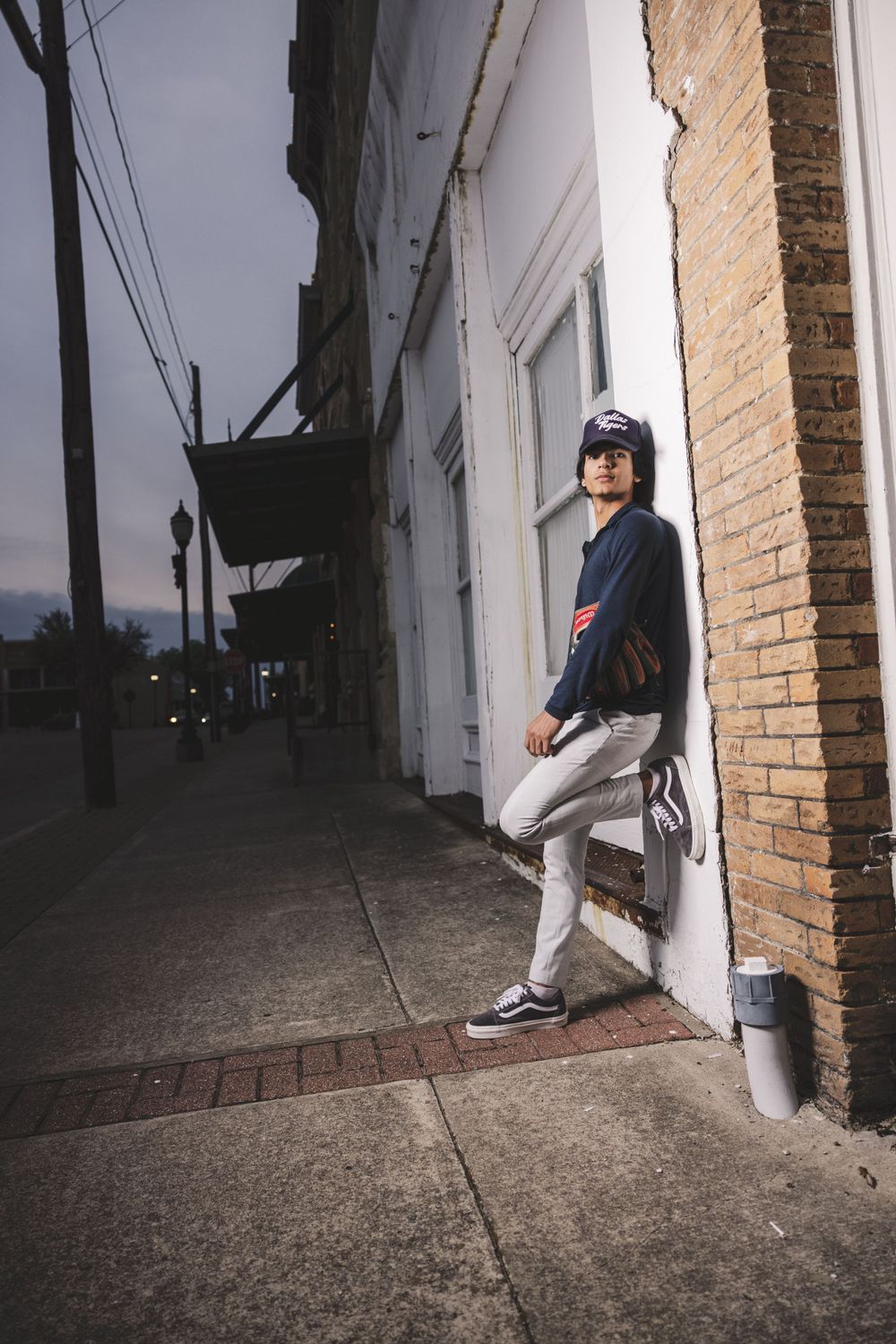 Baseball player in uniform leans against brick building at dusk in urban setting.