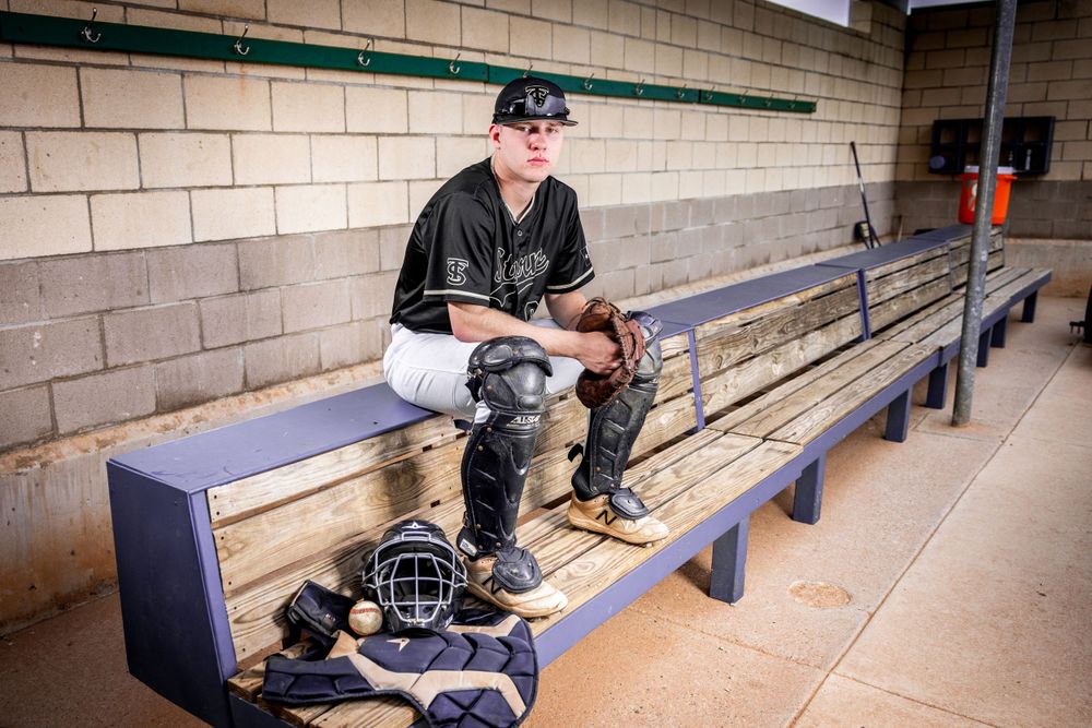 Baseball player sitting in dugout with catcher's gear and equipment.