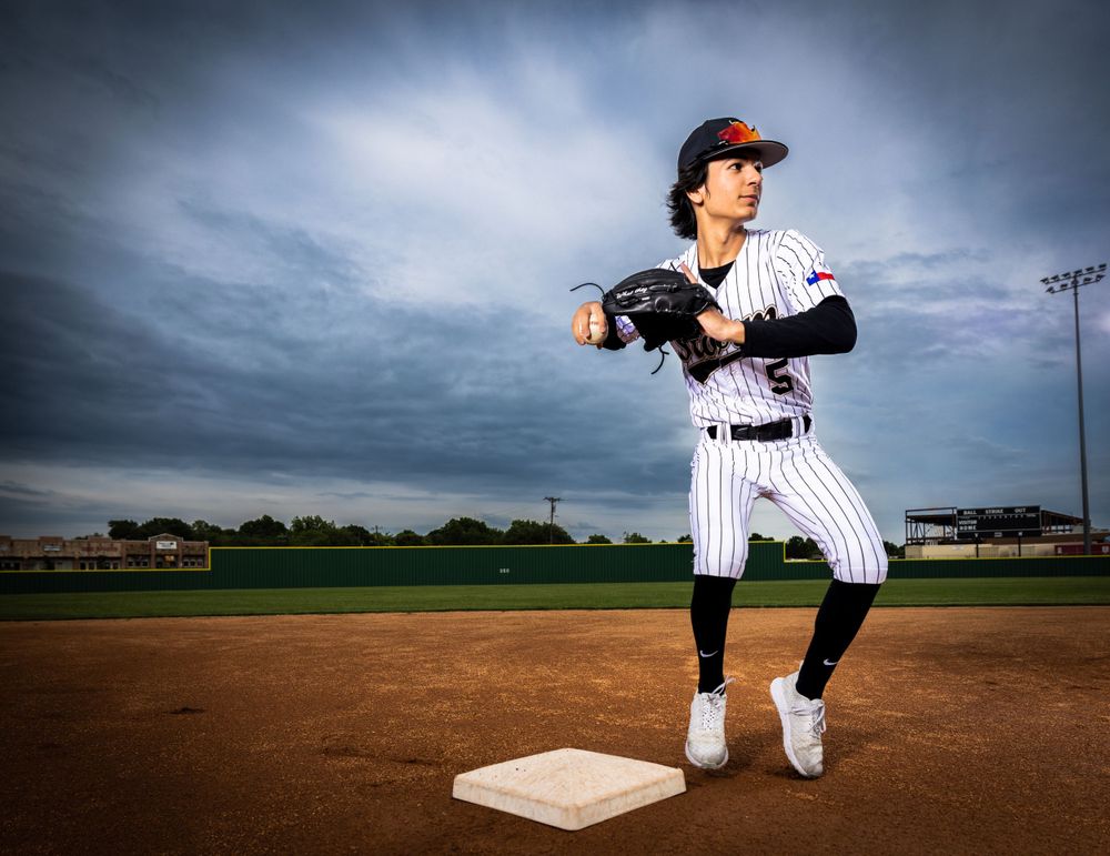 Baseball player in pinstripes winds up for a pitch on a baseball field at dusk with dramatic cloudy sky.