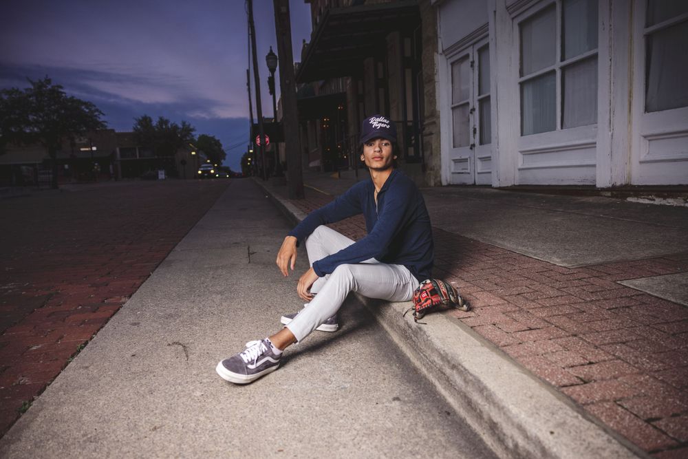 Baseball player in uniform sits on curb along urban street at twilight.