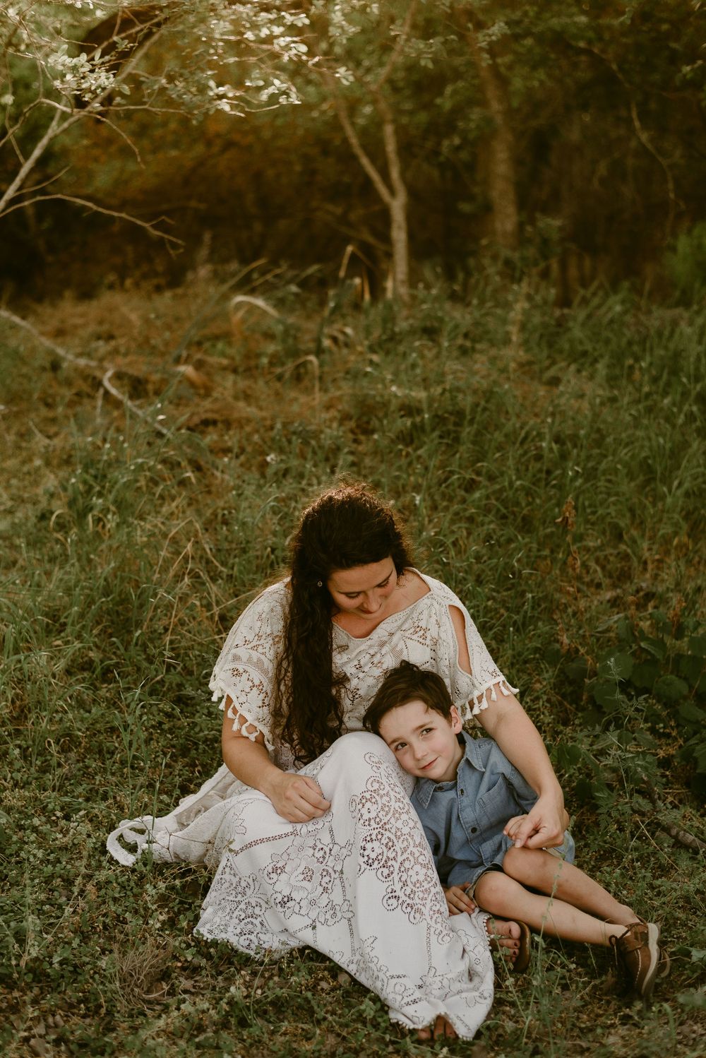 Mother in white lace dress embraces child on grassy hillside during sunset.