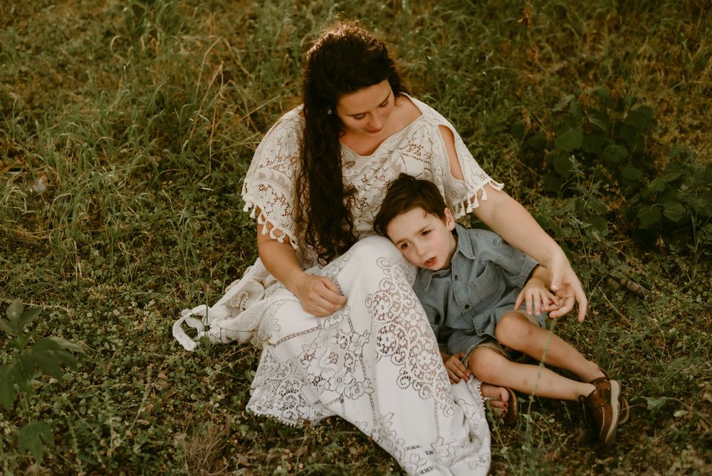 Mother and child share tender moment while sitting on grass in white lace dress.
