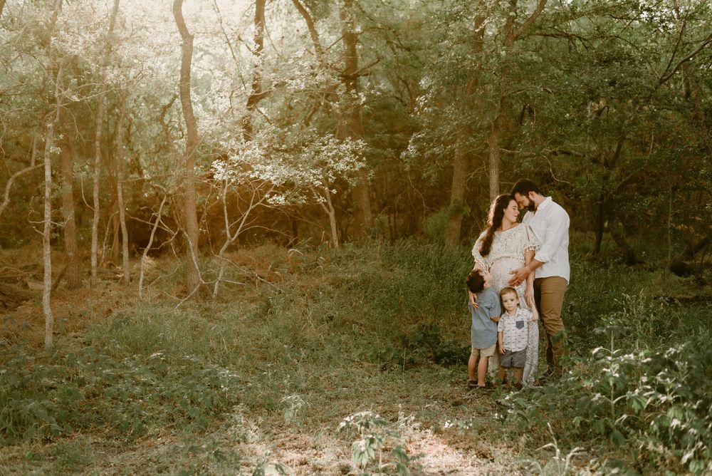 Family shares an intimate moment together in a sunlit wooded area wearing coordinated neutral outfits.