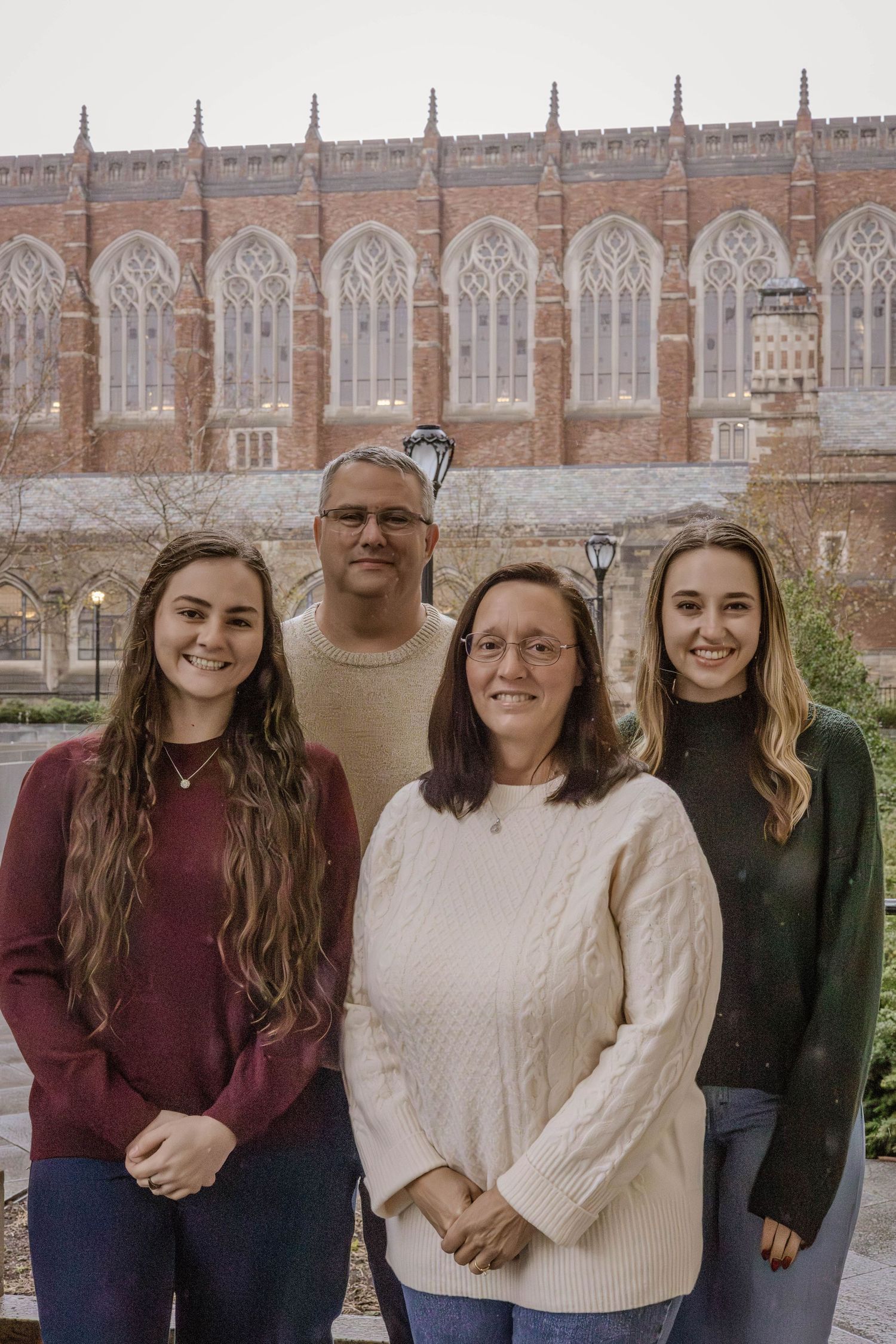 Family of four poses together in front of gothic university building with arched windows.