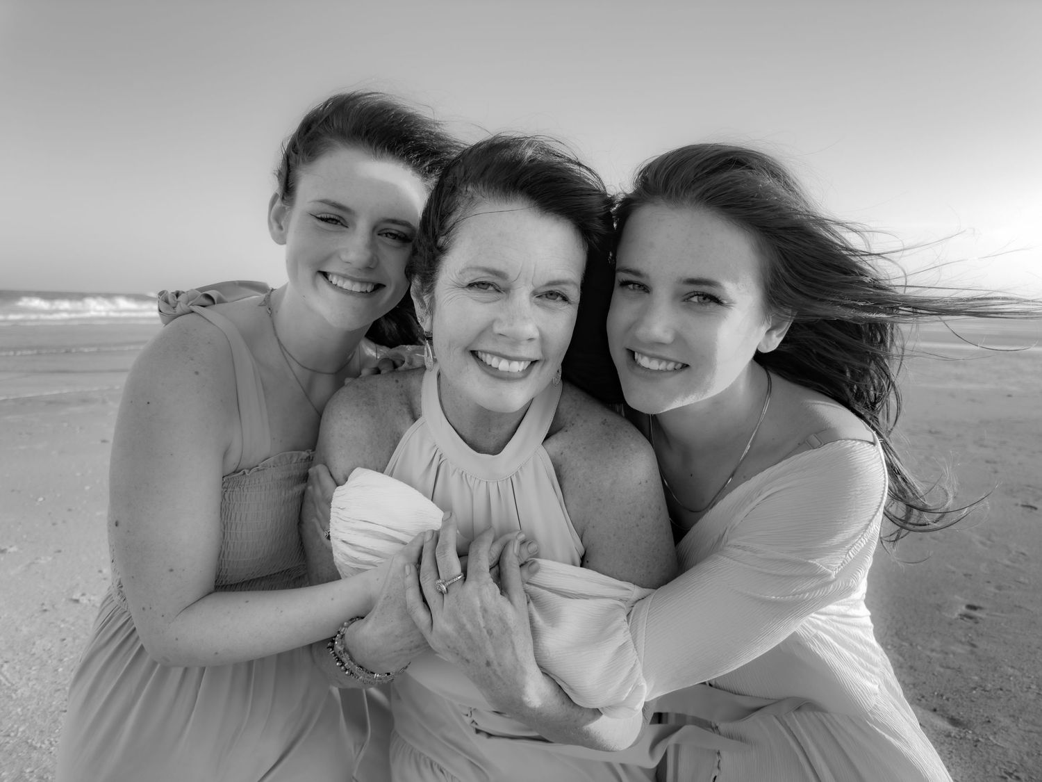 Three women share a joyful embrace on a windy beach in black and white.