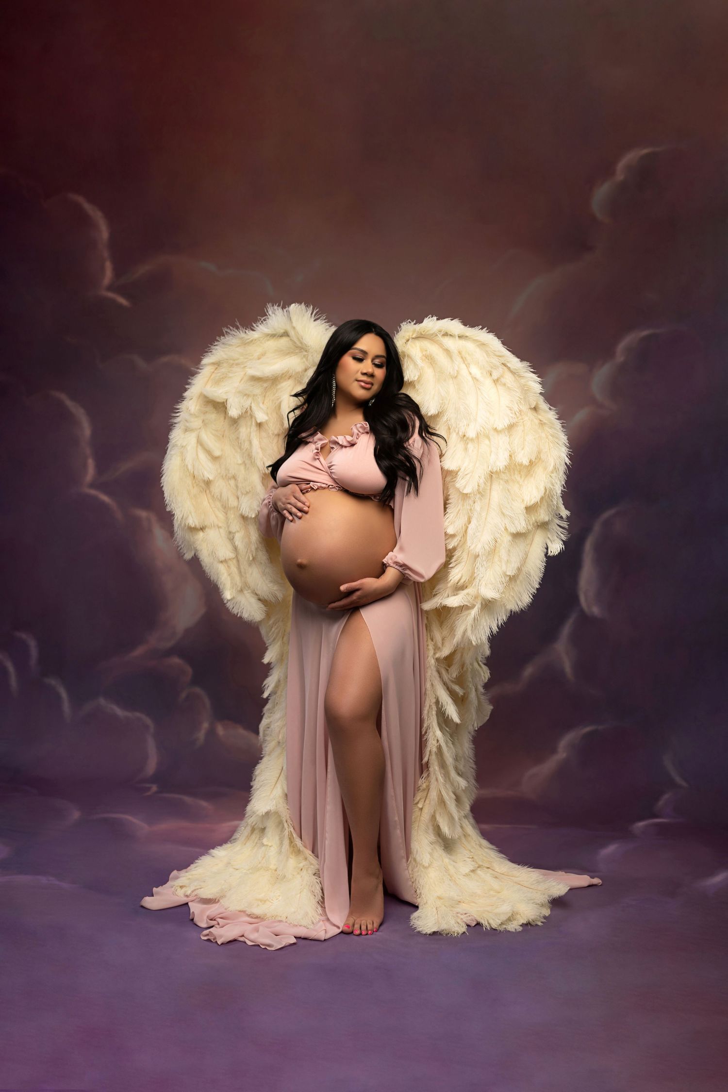 Ethereal maternity photo featuring a flowing blush robe and large white angel wings against a dramatic purple clouded backdrop.