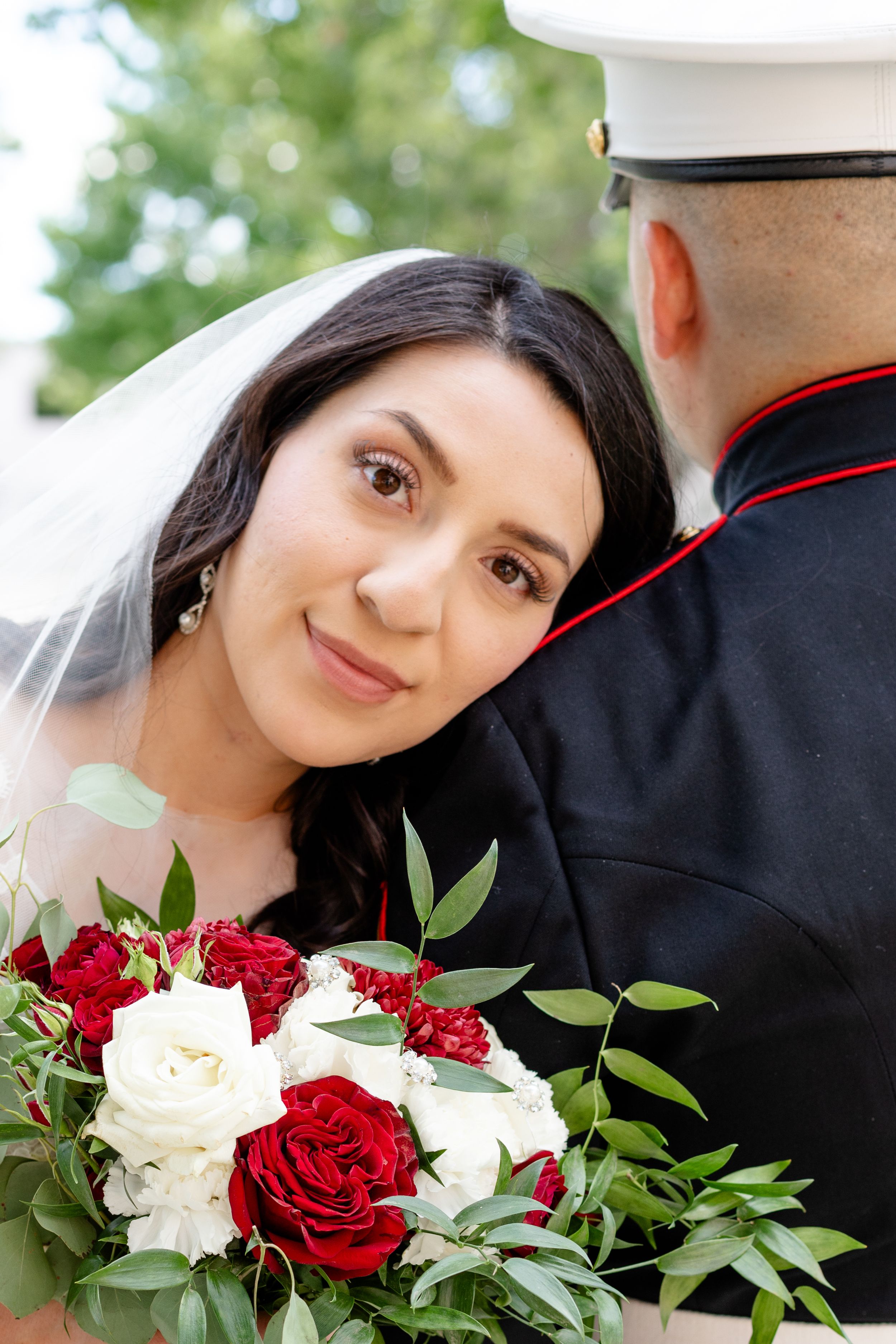 A bride holding a beautiful bouquet of red and white roses with greenery during an outdoor military wedding ceremony.