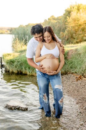 Expecting couple shares intimate moment by waterfront during golden hour.