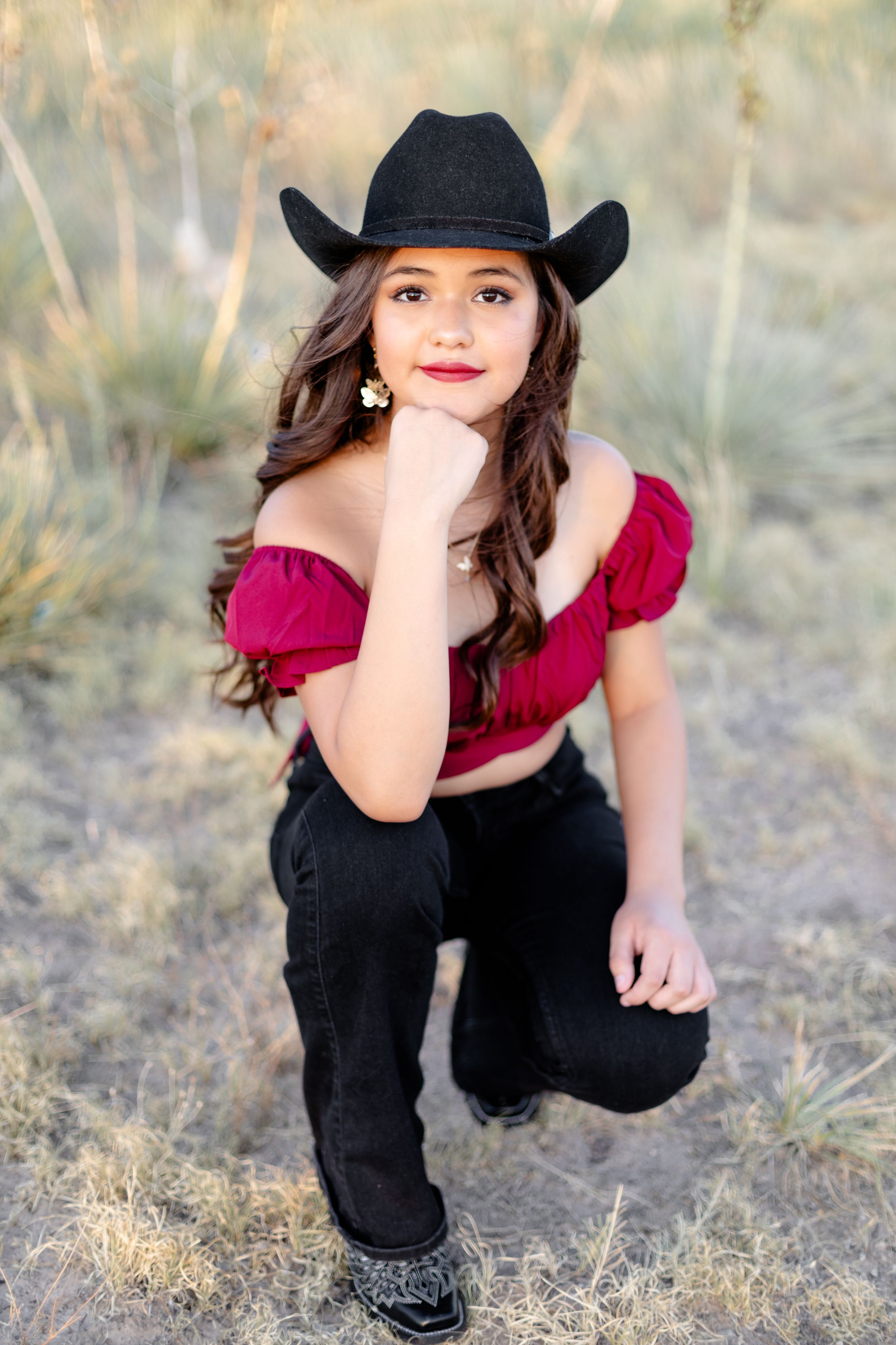 Young woman in red off-shoulder top and black cowboy hat posing outdoors in rustic setting.