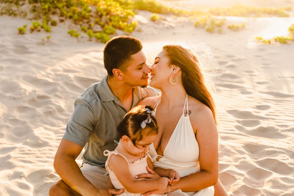 Young family sitting on beach sand at sunset, parents leaning in for a kiss with baby on lap.