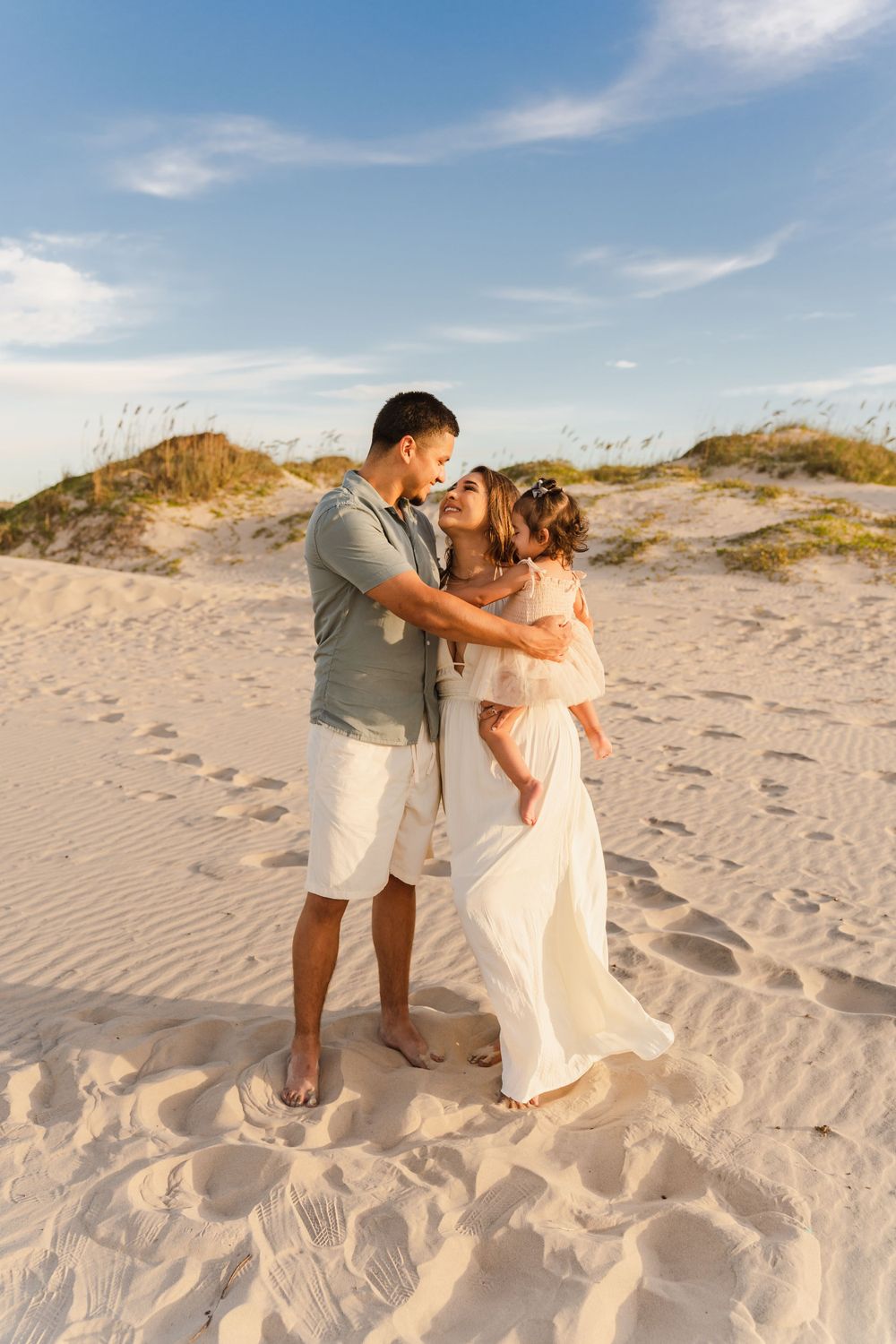 Couple in white outfits holding baby girl share a tender moment on sandy beach dunes at golden hour.