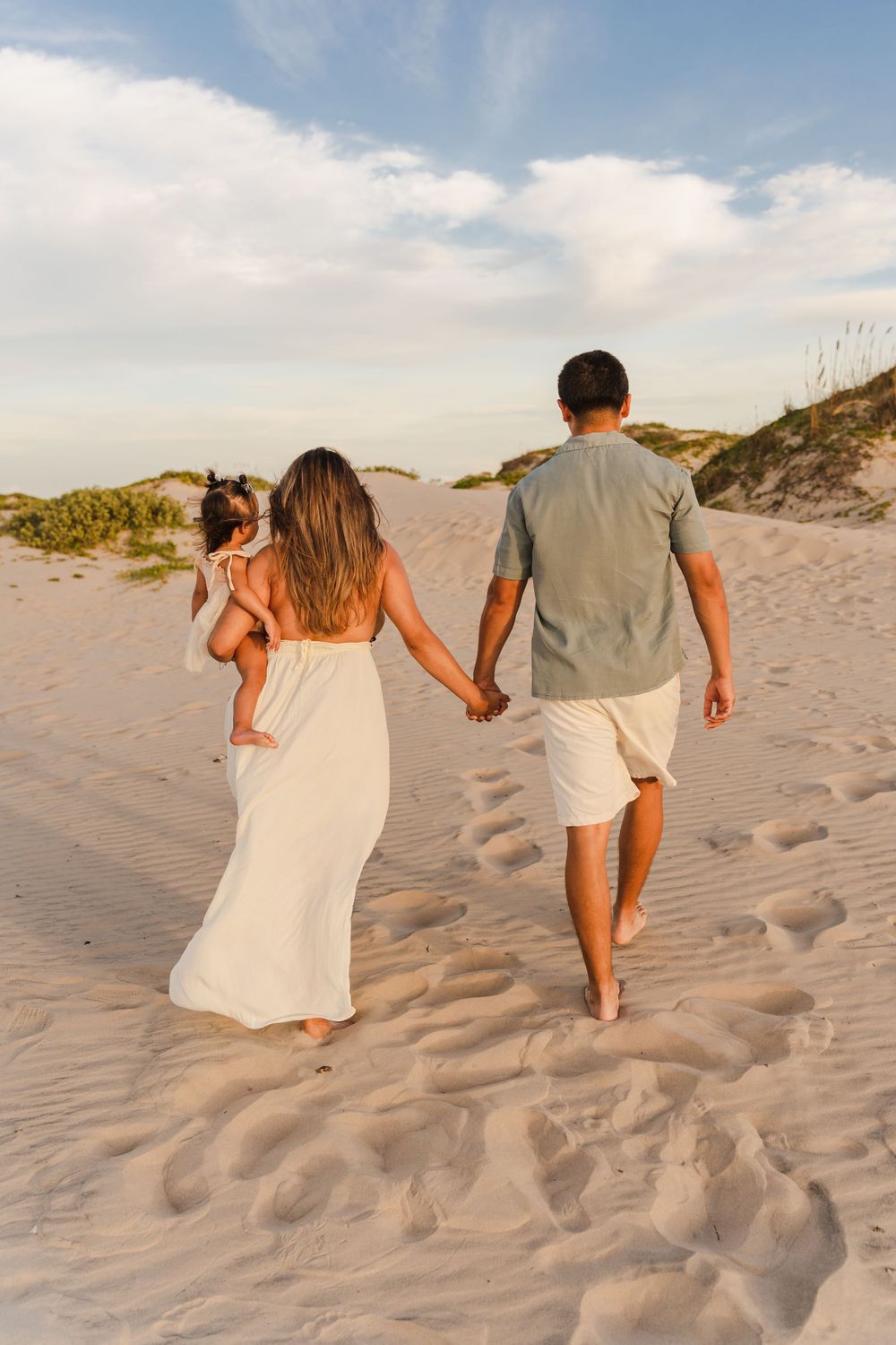 Young family walking away on sandy beach at sunset, mother carrying toddler in white dress, holding hands.