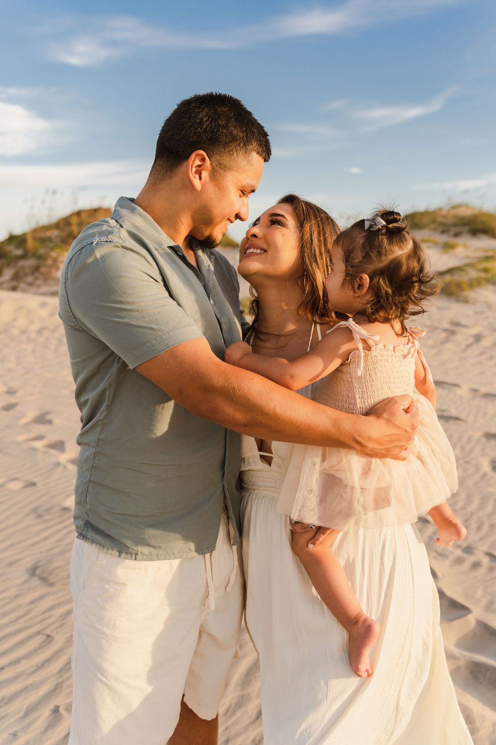 Happy family with father, mother in white dress, and baby girl embracing on sandy beach dunes at sunset.