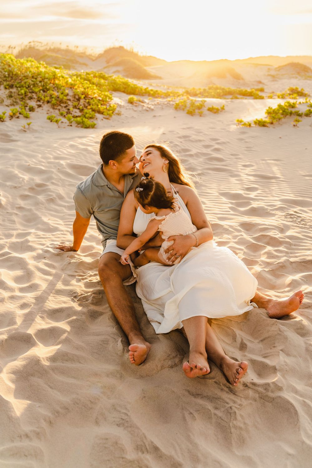 A couple shares a tender moment sitting on sandy beach dunes at golden sunset with their young child.