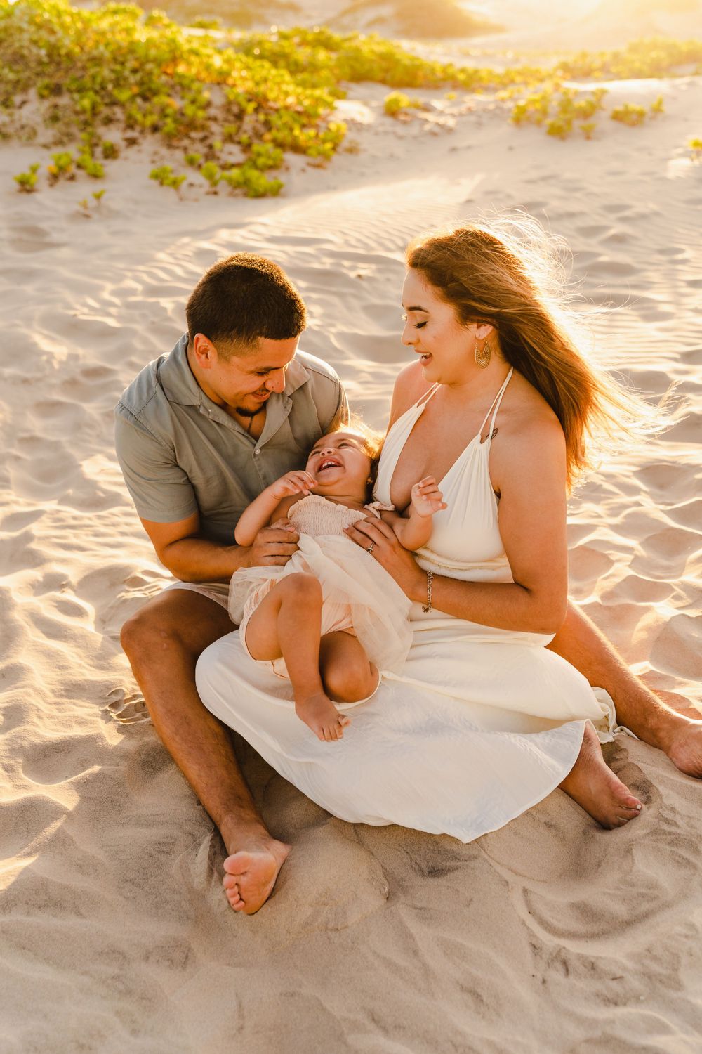 Happy family of three sitting on sandy beach at sunset, parents smiling at laughing baby girl.