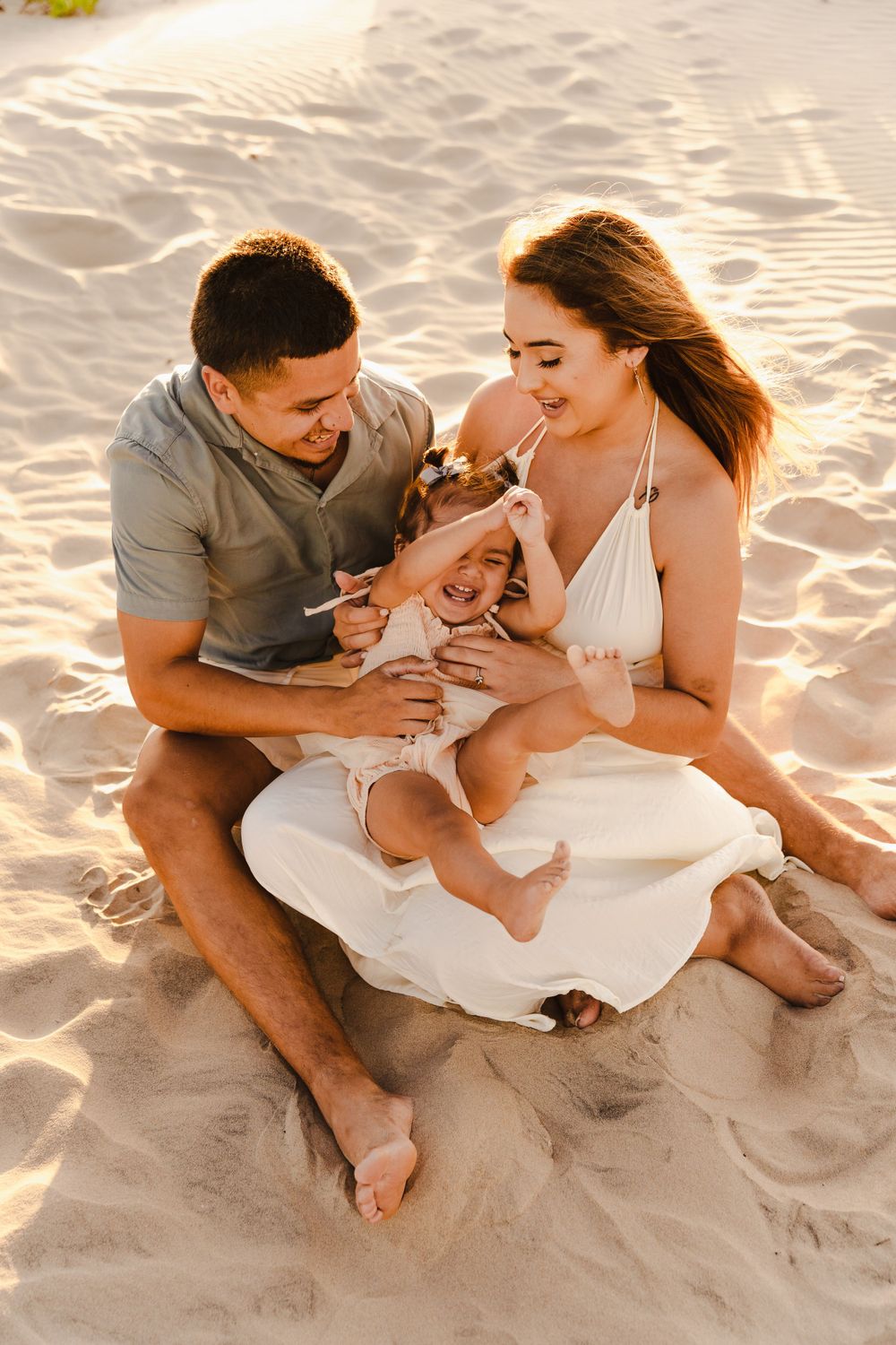 Joyful family sitting on sandy beach at golden hour, parents playing with giggling toddler daughter.