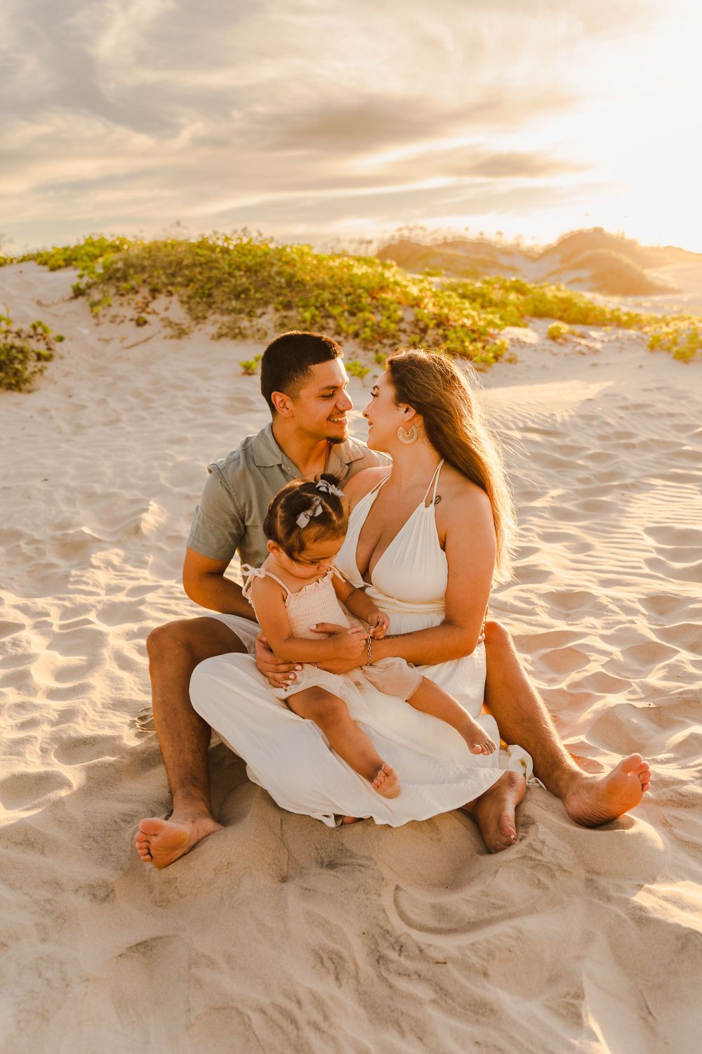 Young couple sitting on sandy beach at sunset, sharing romantic moment while holding their baby girl.