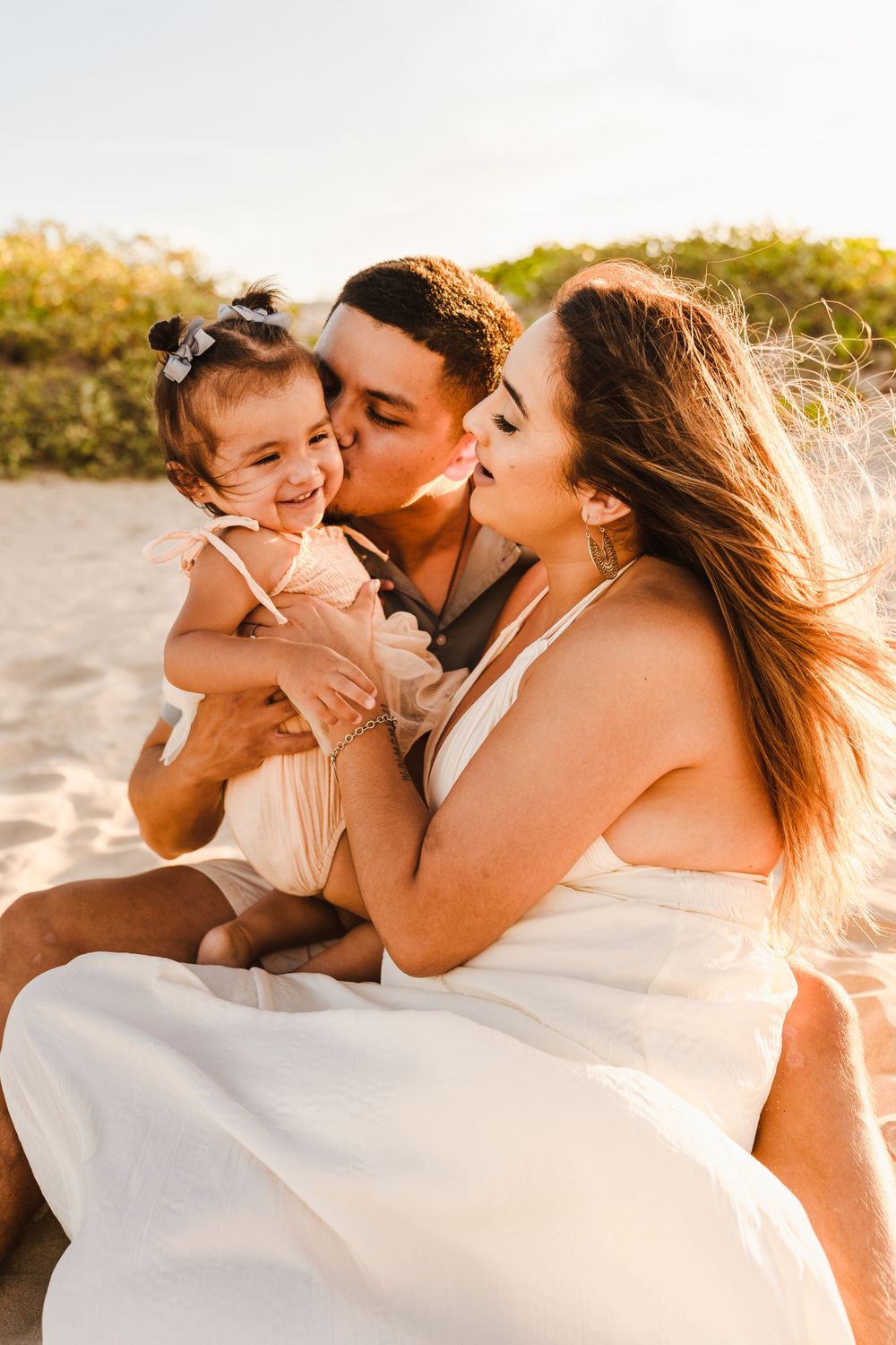 Young couple sitting on sandy beach at sunset, father kissing mother while holding their smiling baby girl.