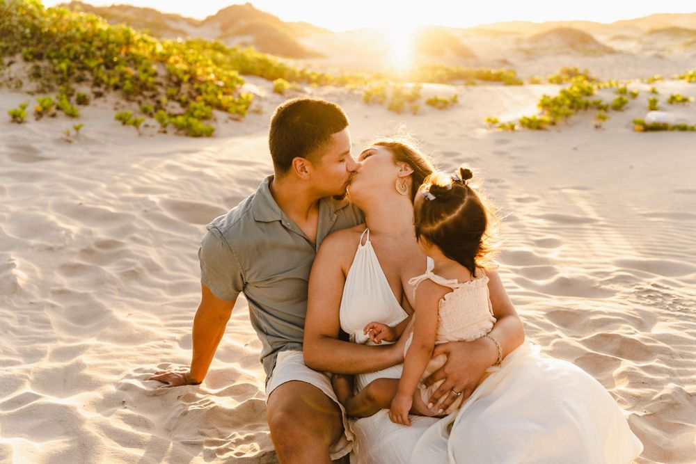 A couple shares a romantic kiss while sitting on white sand dunes bathed in warm golden sunset light.