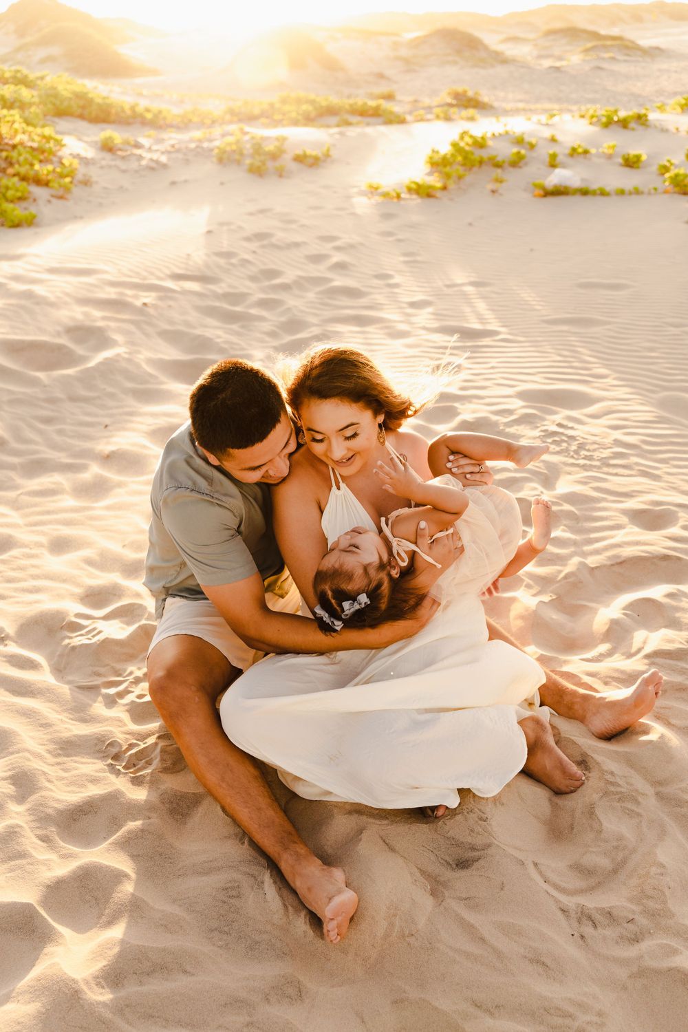 Loving family cuddling together on sandy beach at golden sunset, parents embracing laughing toddler girl.