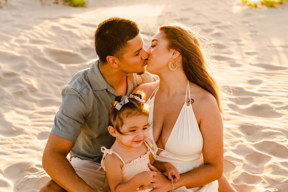Couple sharing a kiss on sandy beach at golden hour while their toddler daughter sits between them.