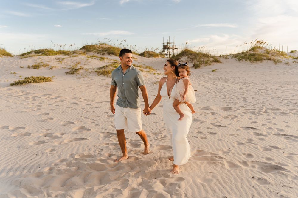 Happy family of three walking barefoot on sandy beach dunes at golden hour, woman in white dress holding hands.