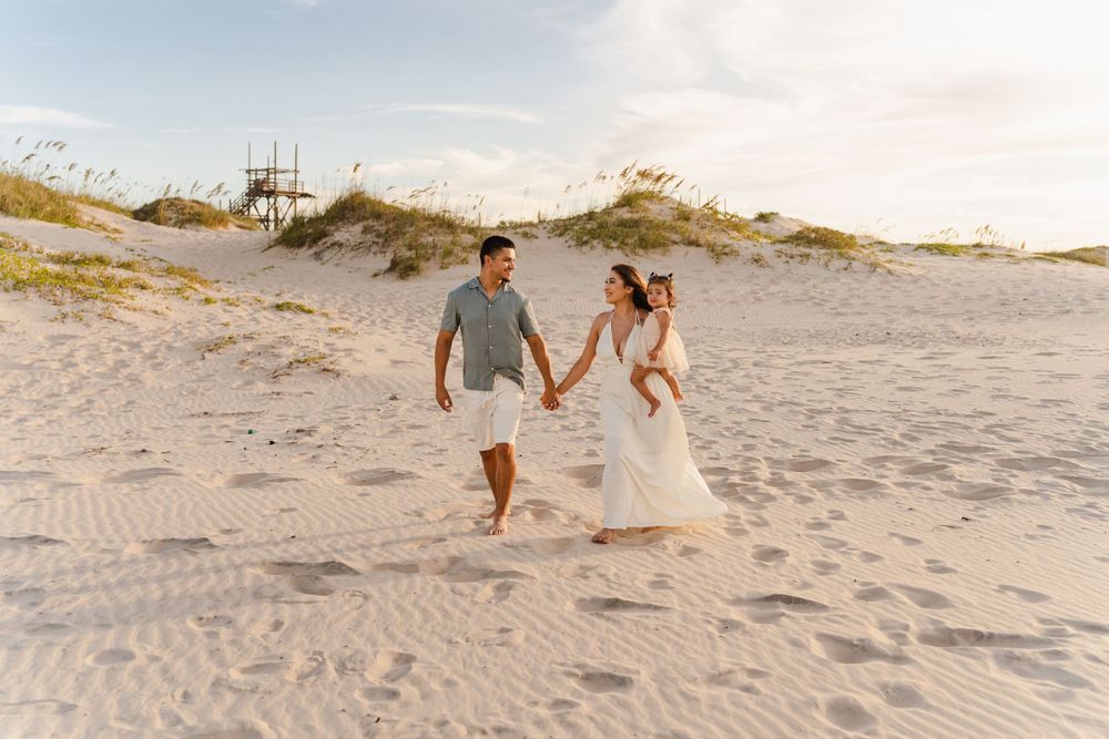 Joyful couple holding hands and walking barefoot on sandy beach dunes with baby in white at sunset.