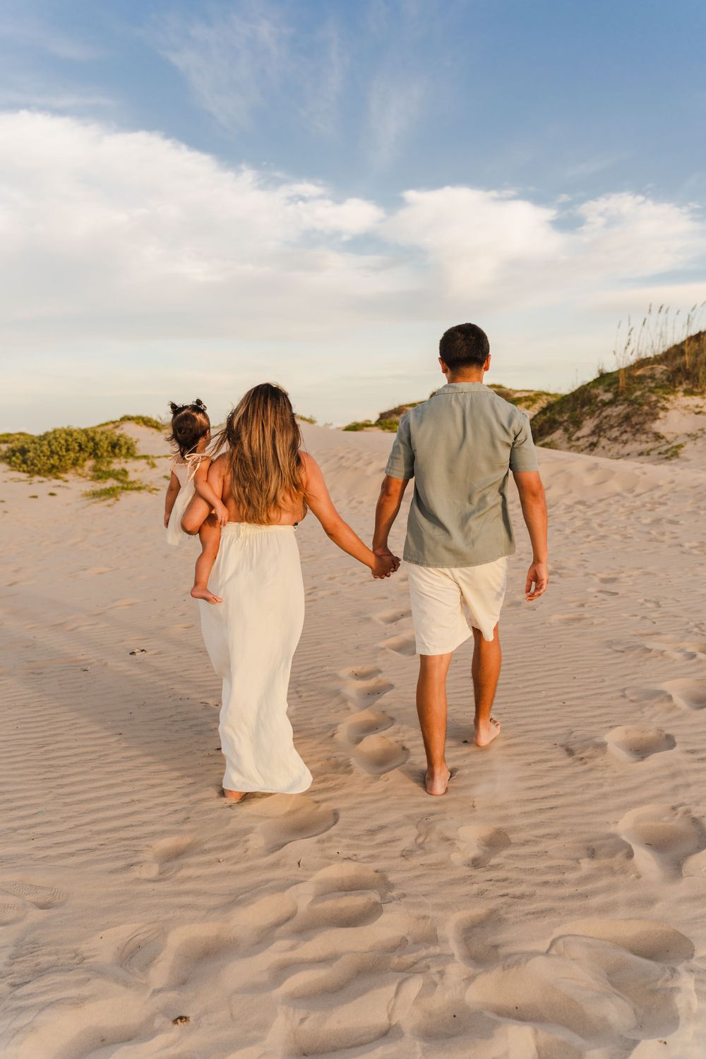 Rear view of young family holding hands walking toward sand dunes at golden hour beach sunset.