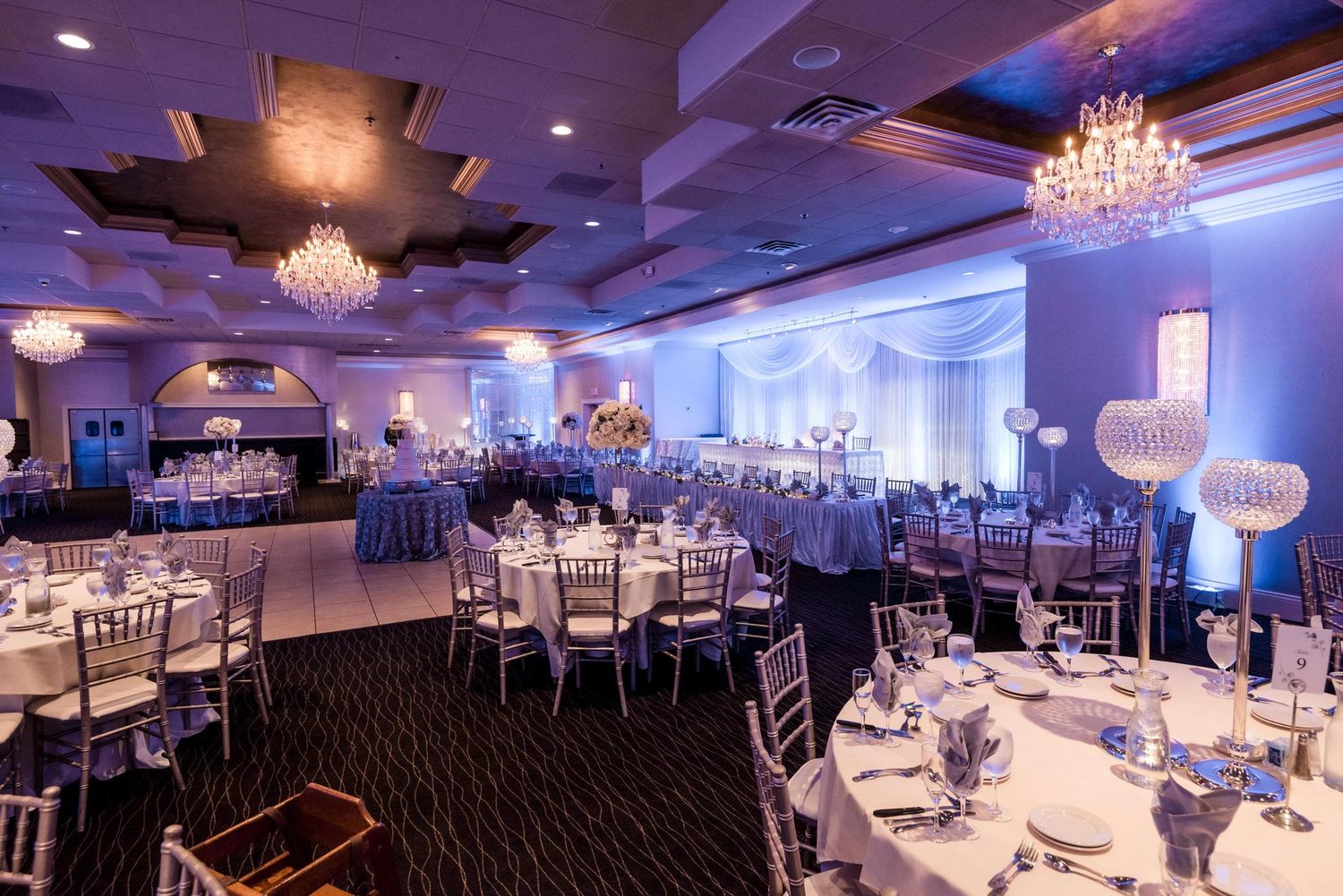 Elegant ballroom setup with crystal chandeliers and formal table settings in blue lighting.