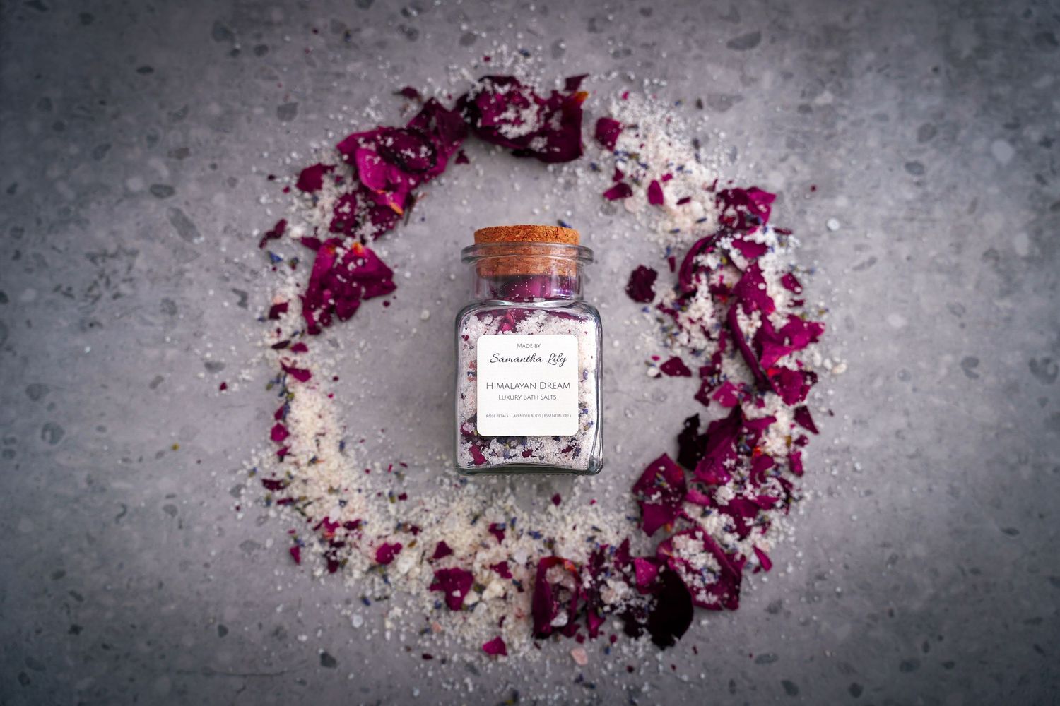 Dried purple flower petals scattered around glass jars with metal lids on textured gray surface.