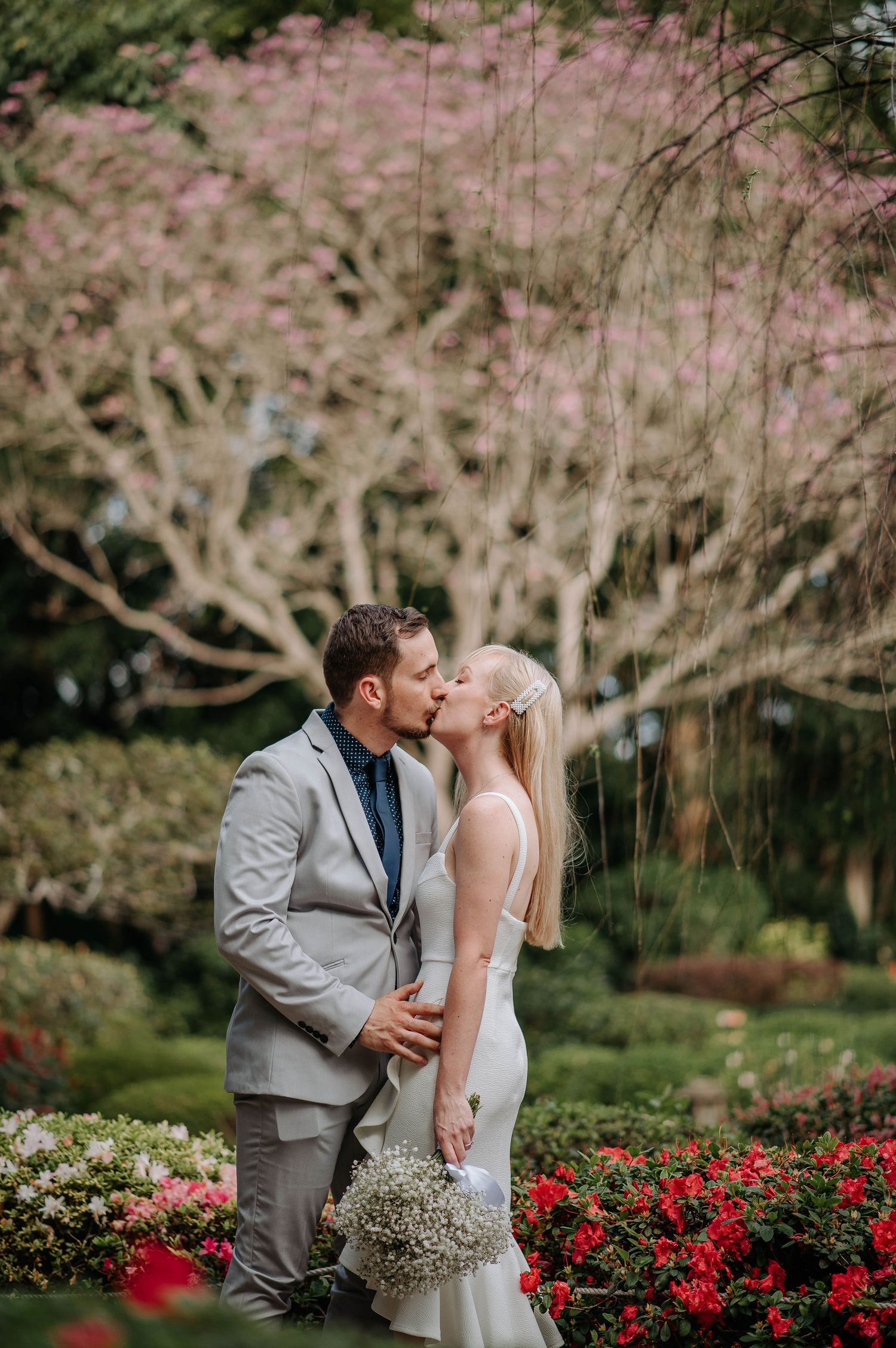 Intimate wedding portrait with couple kissing among cherry blossoms and red garden flowers.