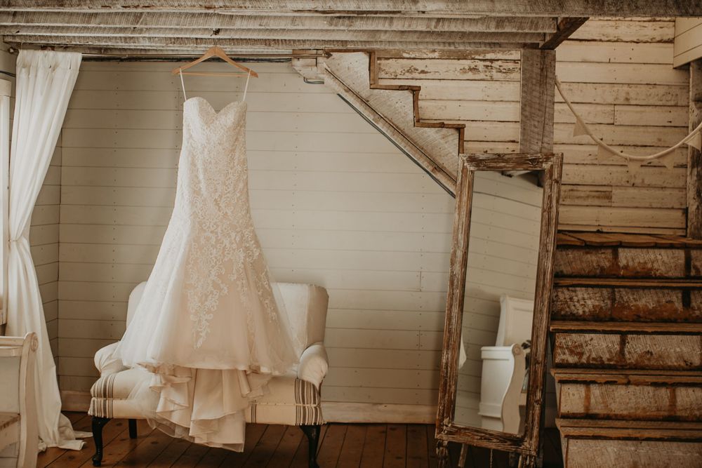 Lace mermaid wedding gown hanging near vintage mirror and staircase in charming rustic barn setting.