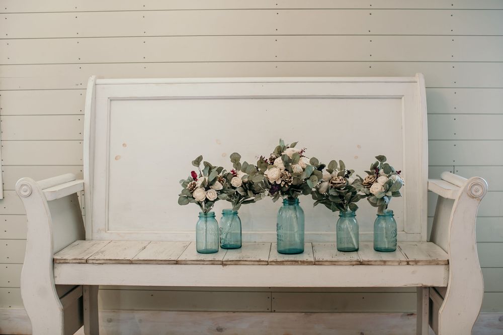 Four blue mason jars with white and pink floral arrangements displayed on rustic white wooden bench.