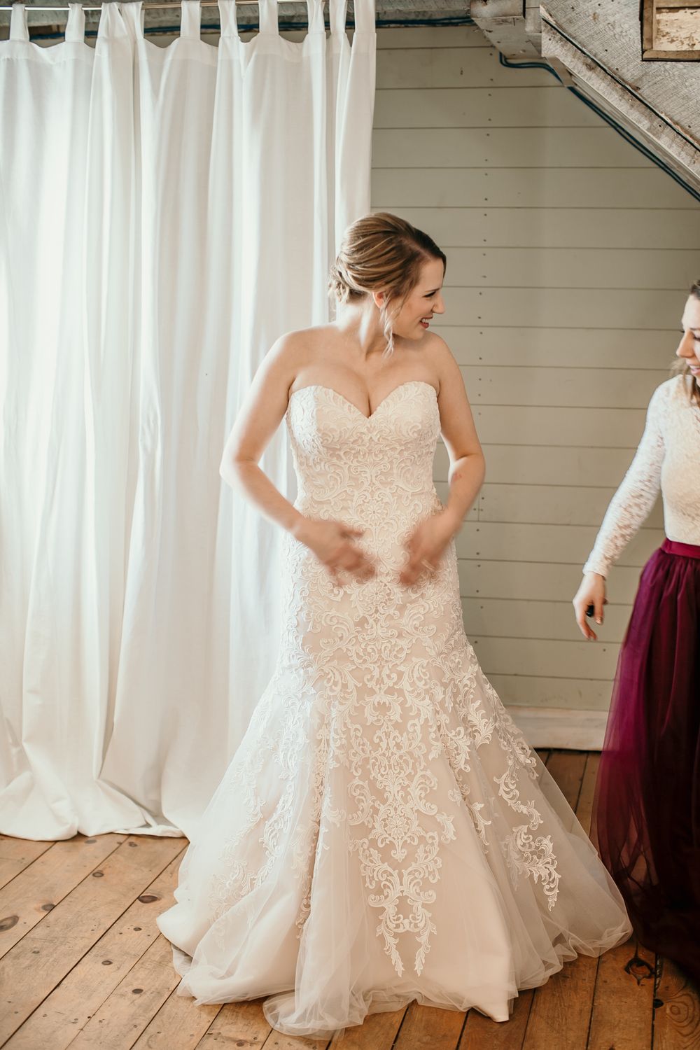 Bride in strapless lace mermaid wedding gown being assisted by bridesmaid in burgundy dress.