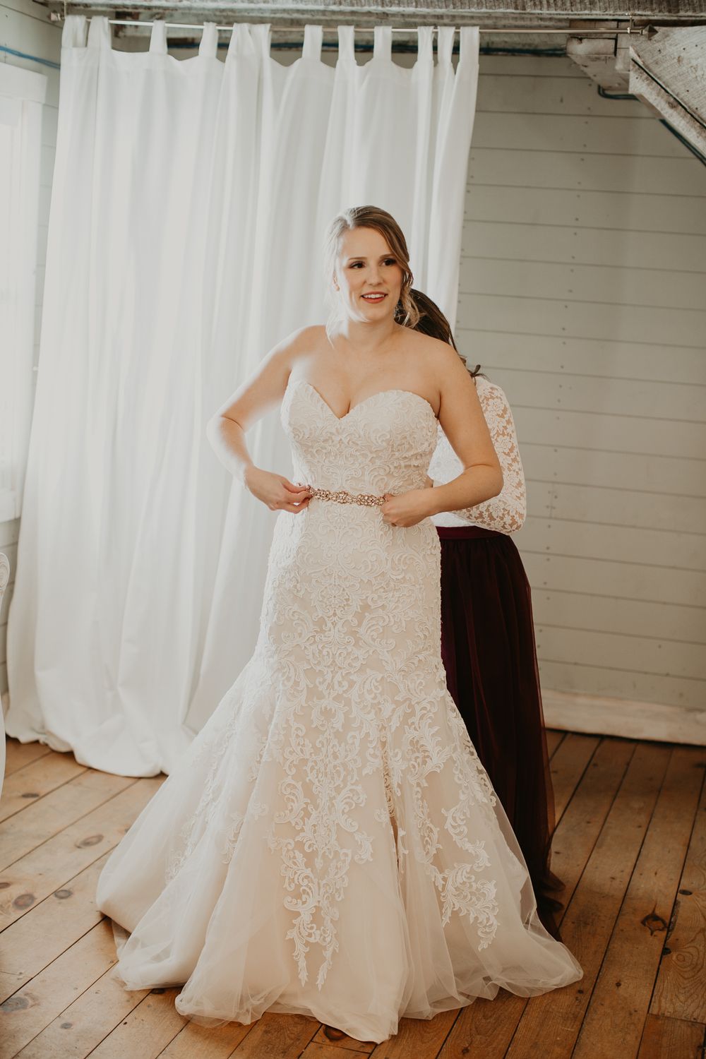 Smiling bride in strapless lace mermaid gown with crystal belt being buttoned by bridesmaid.