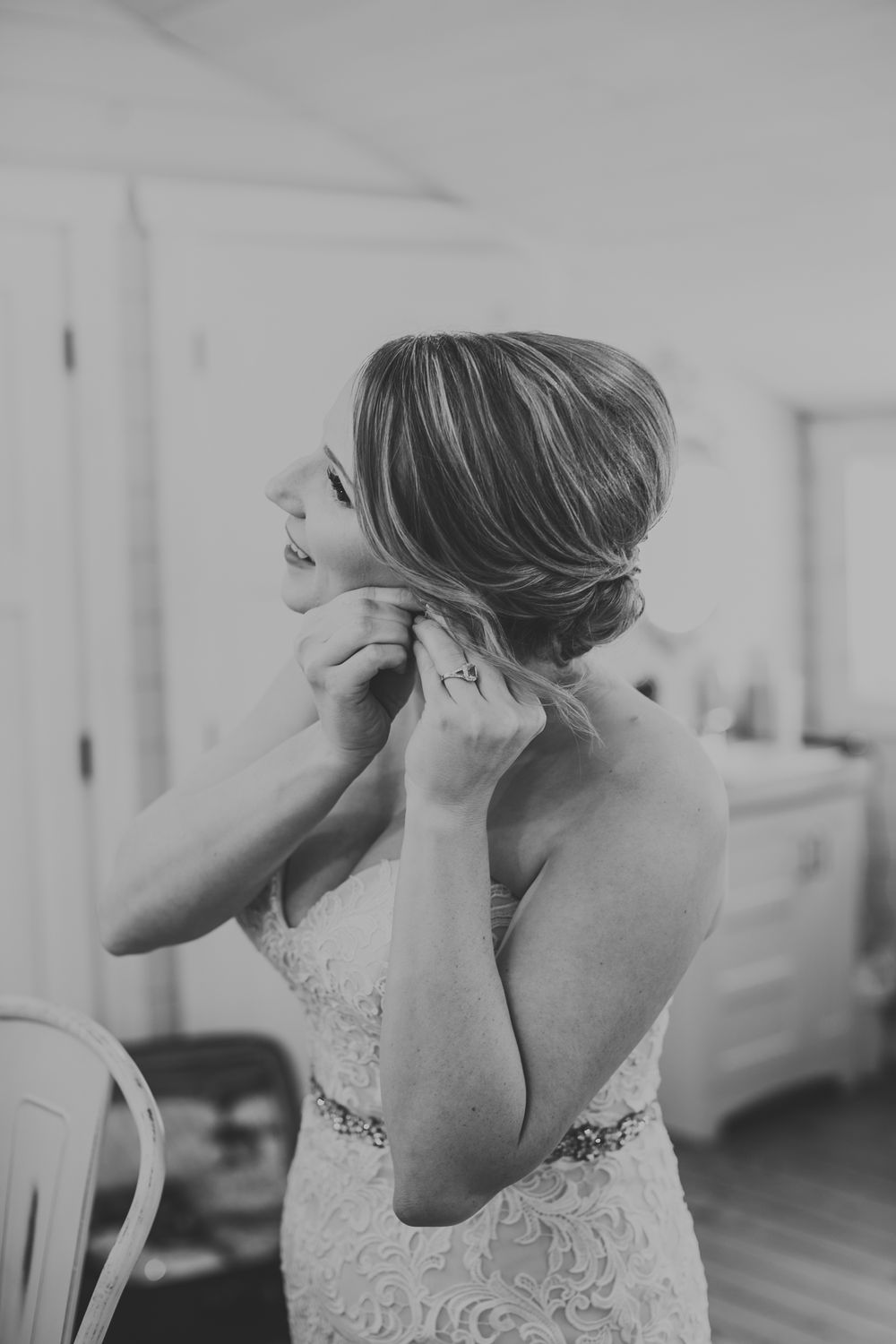 Black and white photo of bride in lace gown putting on earrings with elegant updo hairstyle.