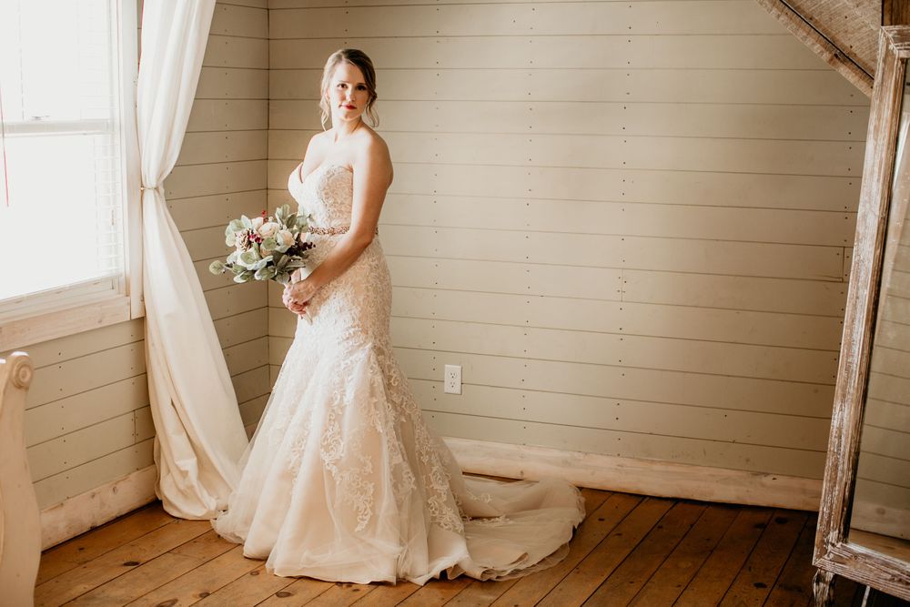 Bride in lace mermaid gown holding bouquet posing by window in rustic shiplap room.